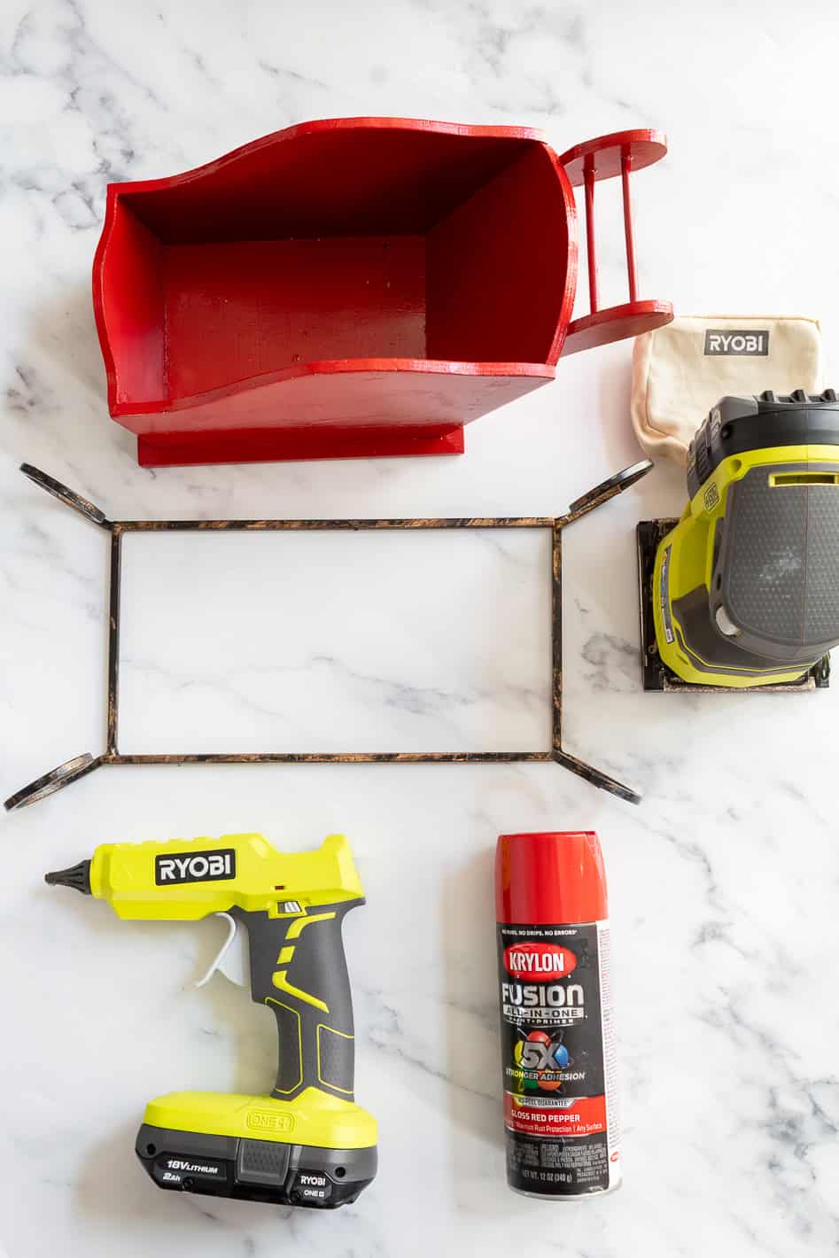 Supplies to make a Christmas sleigh centerpiece including a thrifted sleigh, metal sleigh runner, hand snader, glue gun, and red spray paint