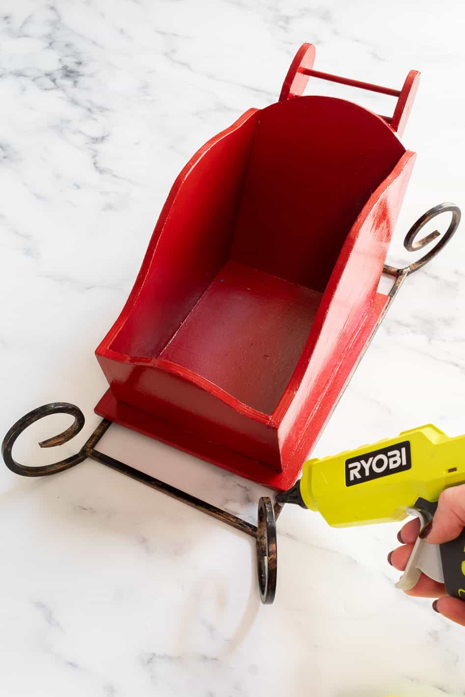 Hot gluing a metal runner onto the bottom of a red wooden sleigh to make a Christmas centerpiece