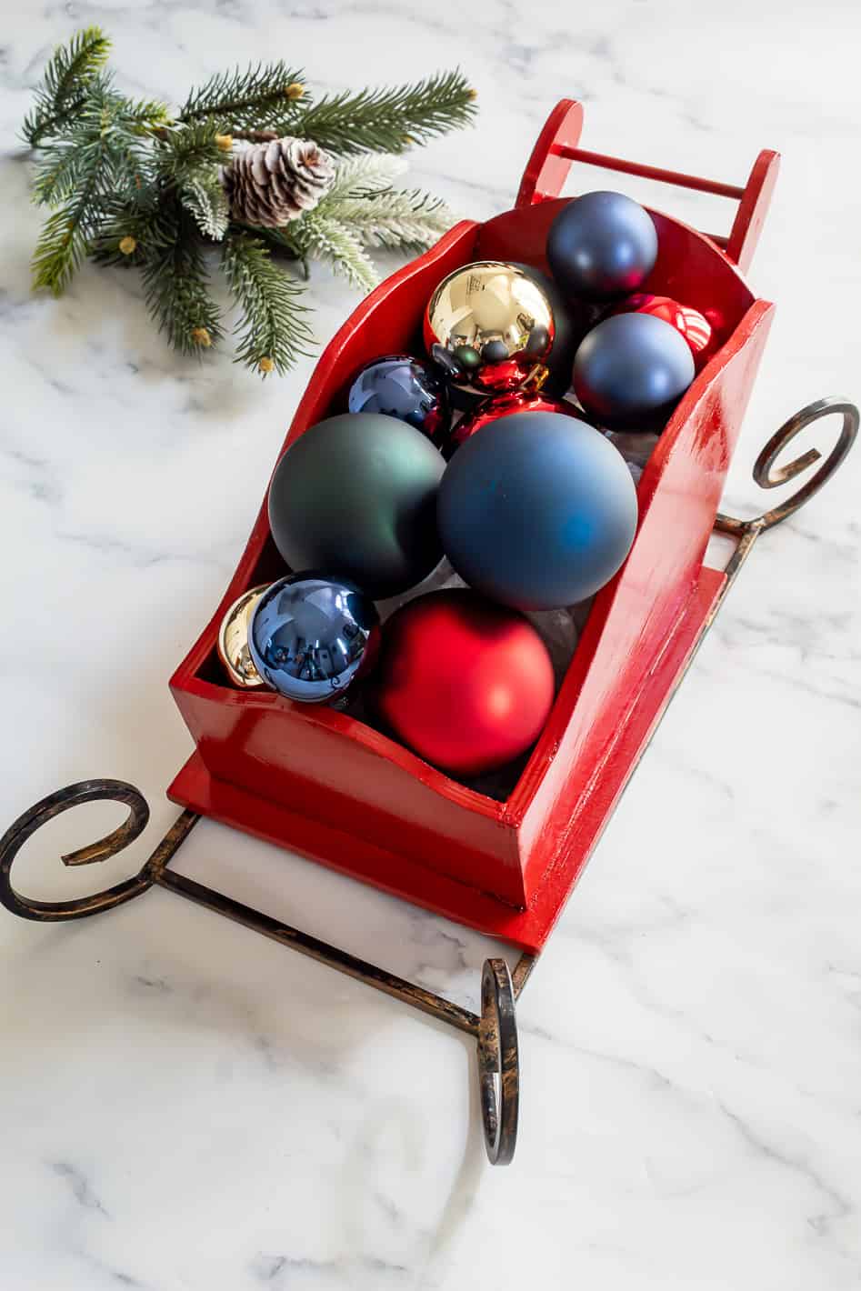 Close up view of faux greenery and red, gold, blue and green ornaments inside of wood sleigh Christmas centerpiece