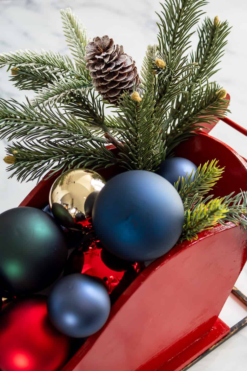 Close up view of faux greenery and red, gold, blue and green ornaments inside of wood sleigh Christmas centerpiece
