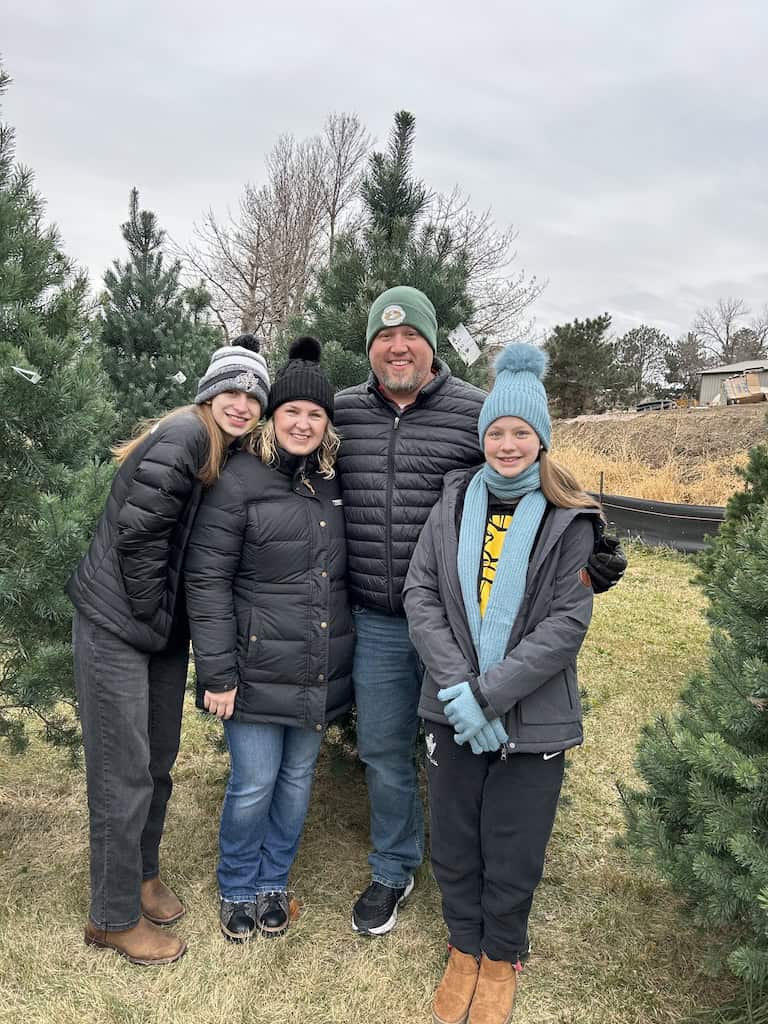 family smiling in front of their Christmas tree on a Christmas tree farm