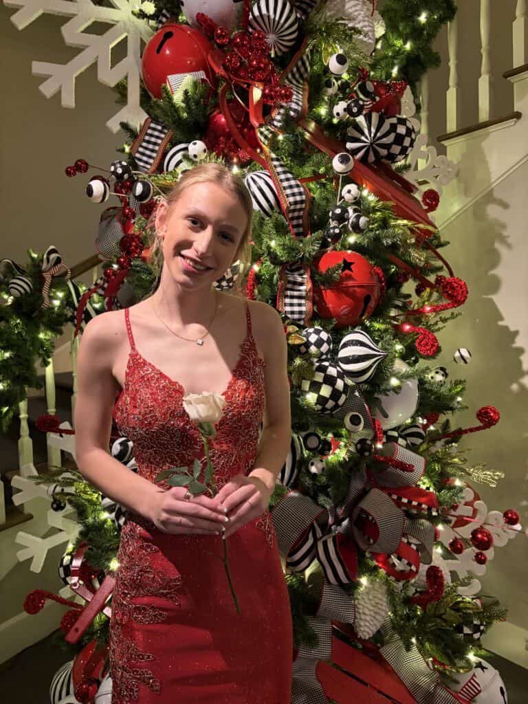girl in a formal dress standign in fornt of a Christmas tree smiling and holding a white rose