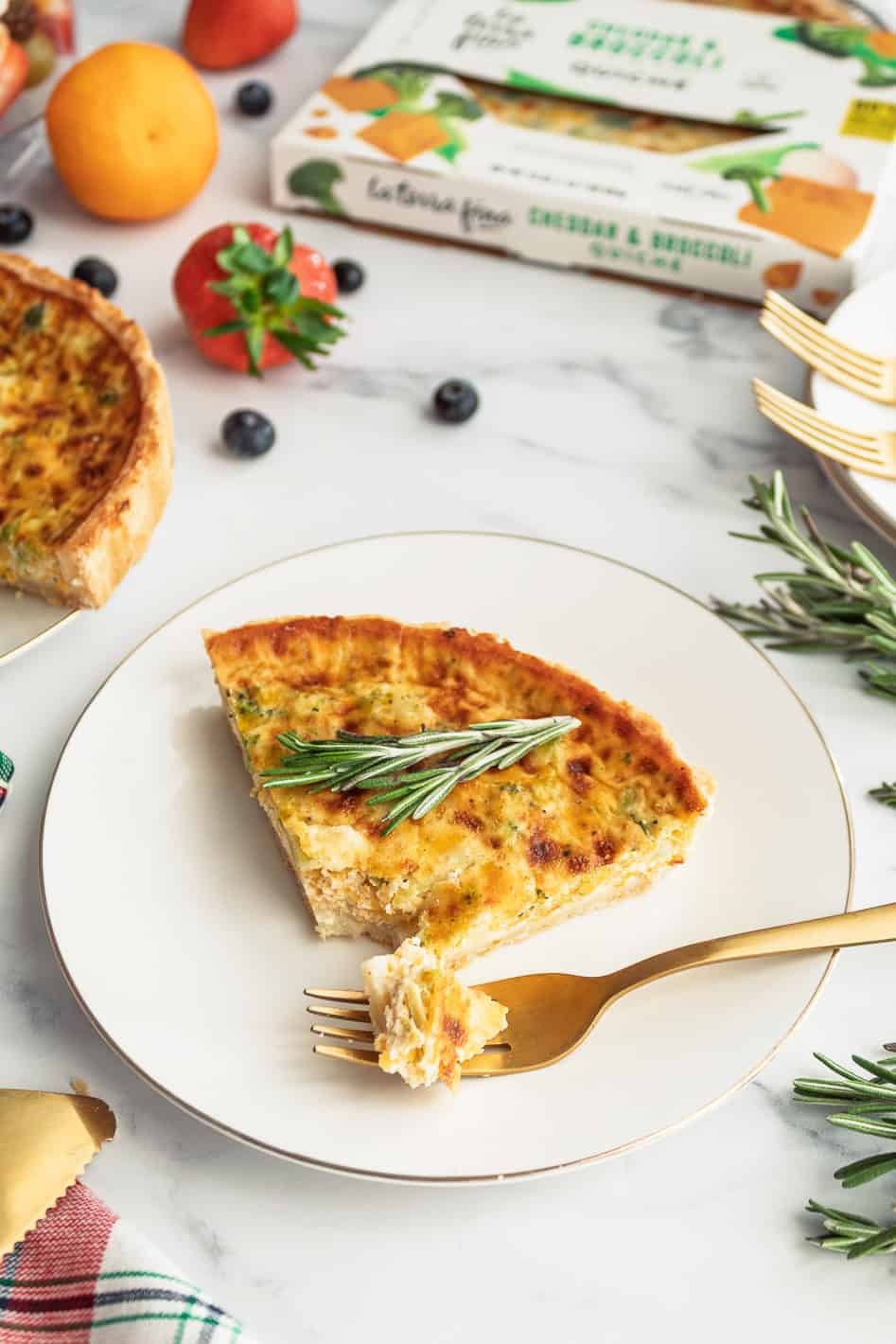 slice of quiche ona plate with a fork and a rosemary garnish with fruit in the background