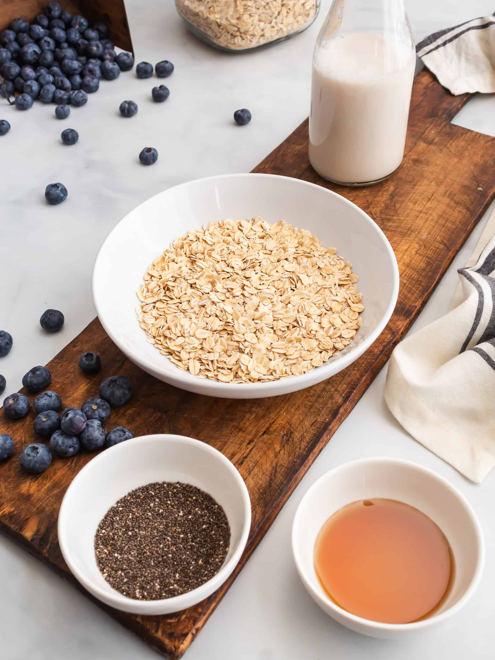 Ingredients for blueberry overnight oats including rolled oats, chia seeds, maple syrup, almond milk, and fresh blueberries arranged on a rustic wooden board.