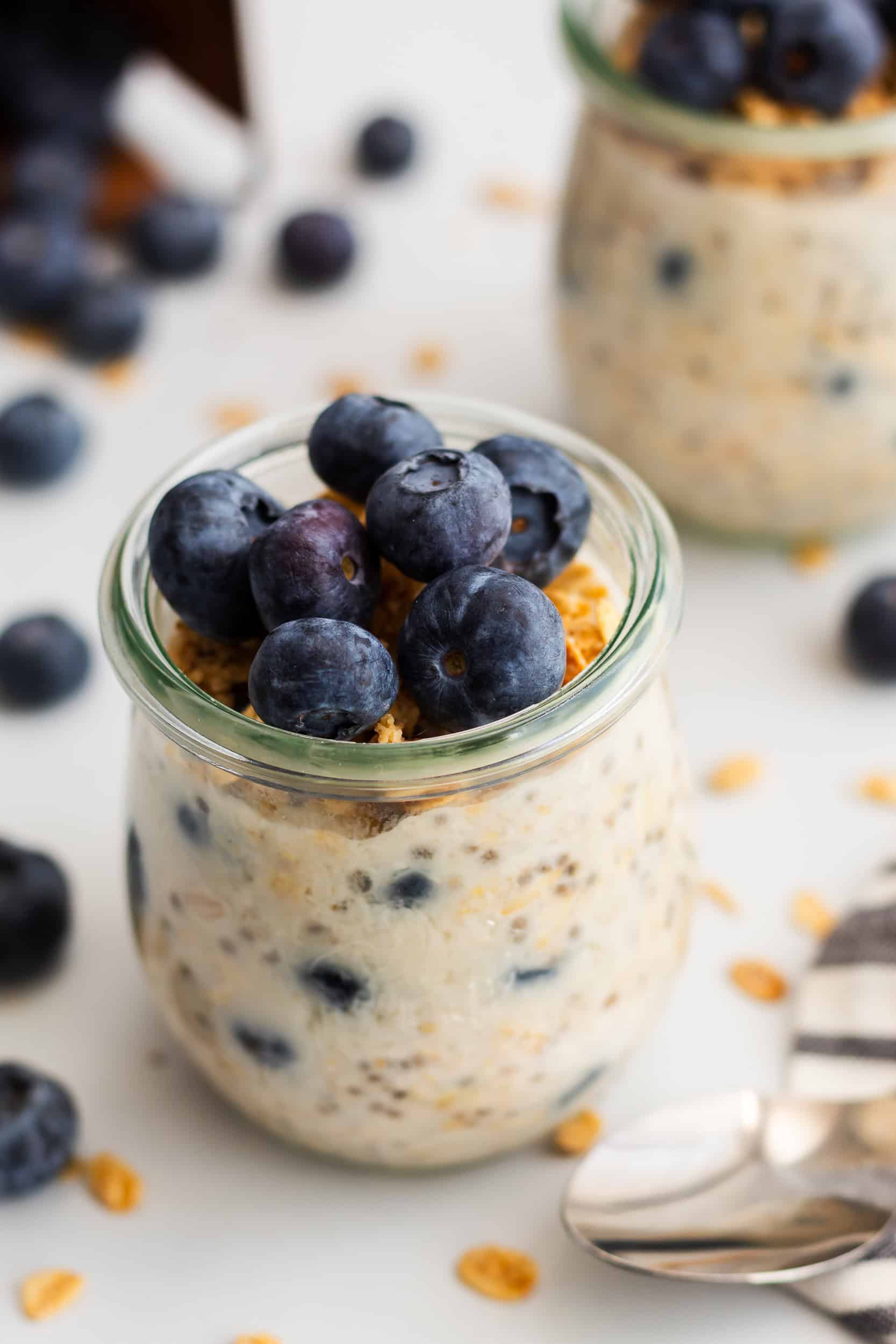 A single serving of blueberry overnight oats in a clear glass jar topped with granola and blueberries, styled with a spoon and scattered oats.