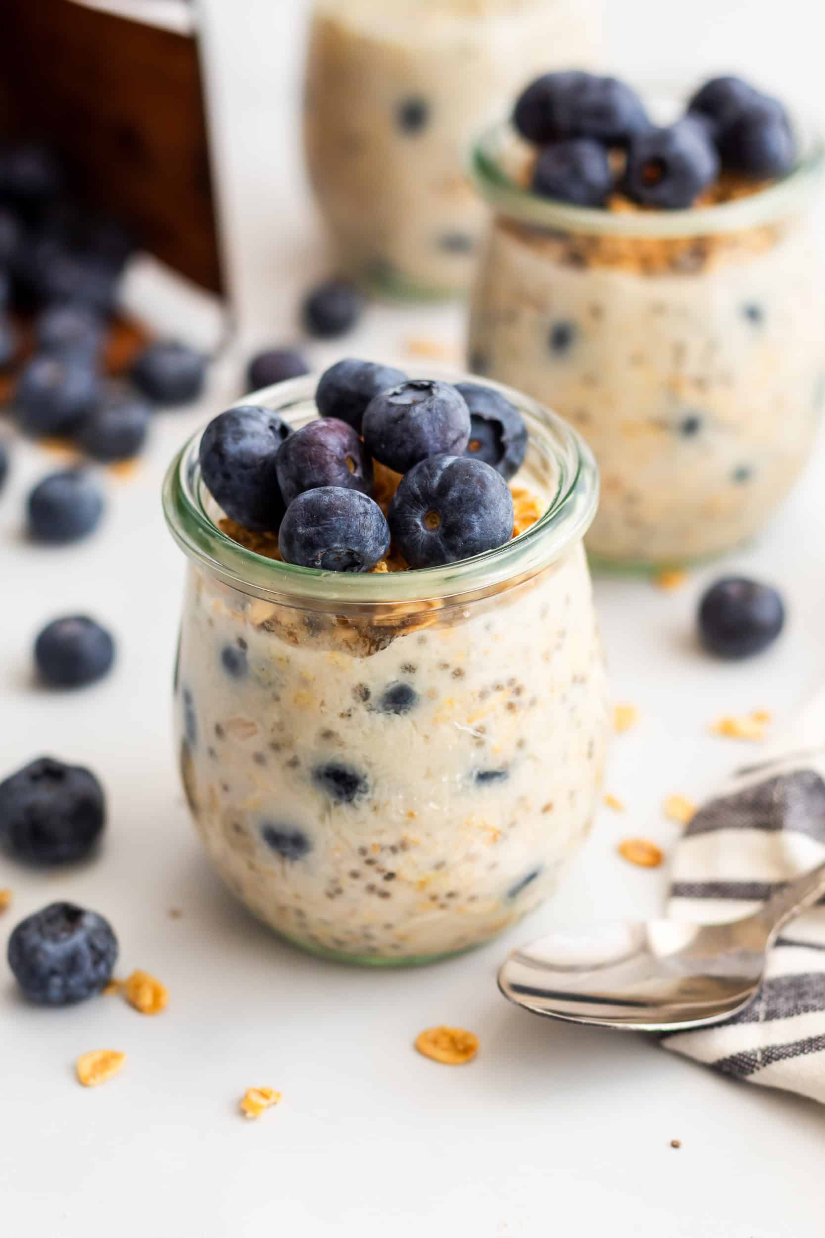 A glass jar filled with blueberry overnight oats topped with blueberries and granola, surrounded by loose berries and oat flakes.