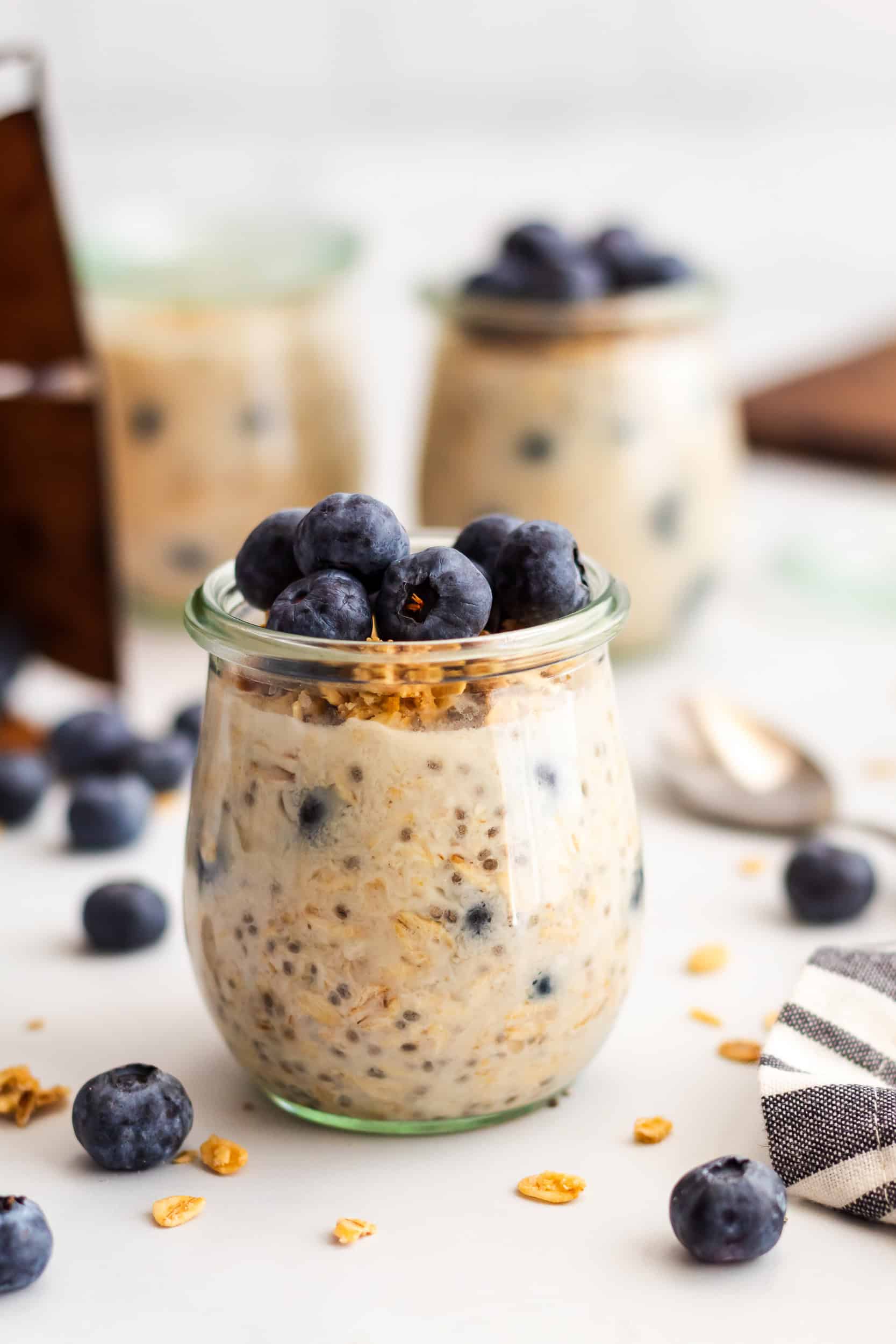 A single glass jar of blueberry overnight oats topped with granola and fresh blueberries, styled with scattered berries and oat flakes.
