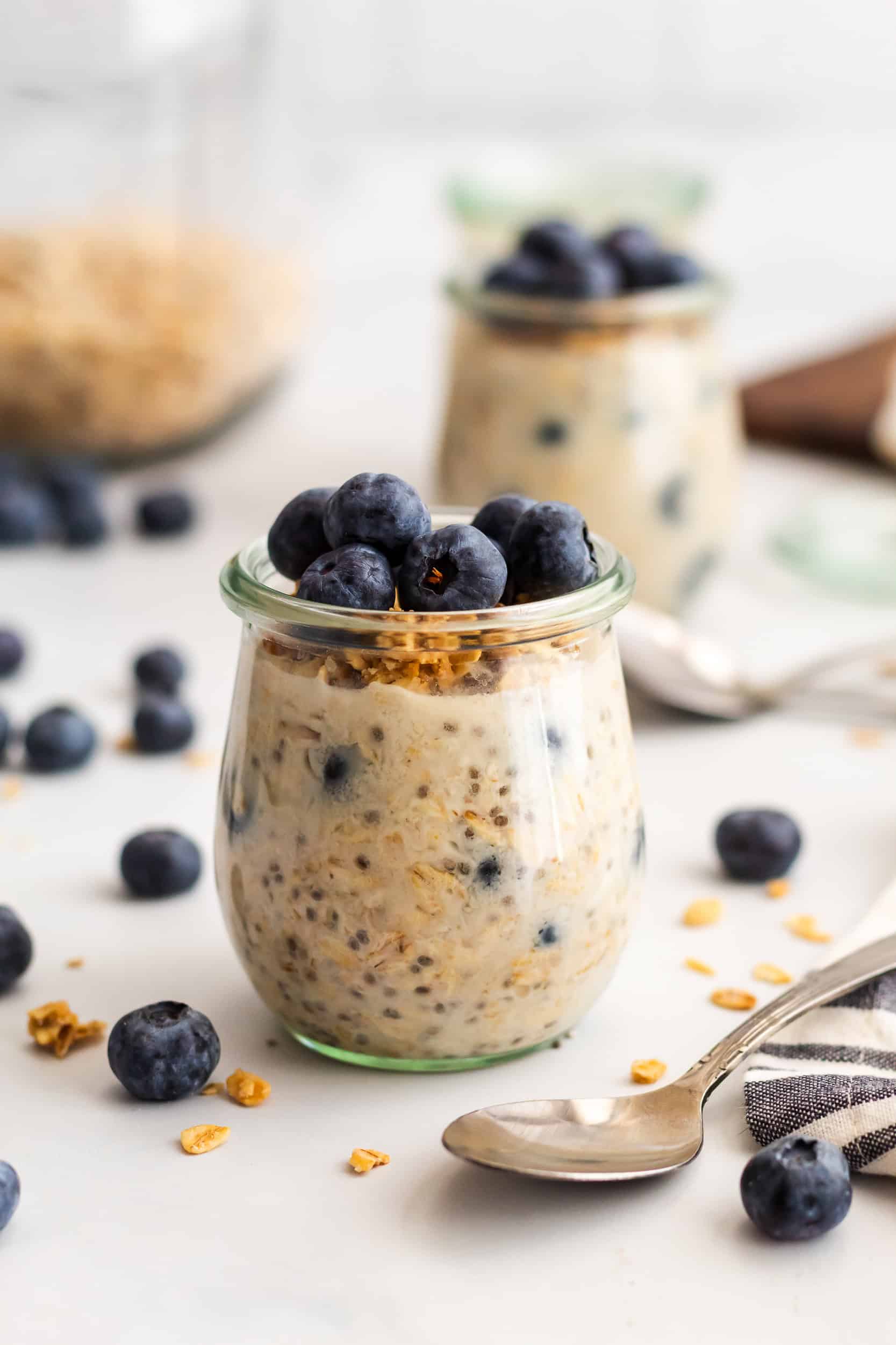 A close up of blueberry overnight oats in a small glass jar with blueberries on top, with a spoon and soft blurred jars in the background.