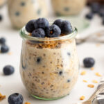 A front facing view of blueberry overnight oats in a clear jar topped with crunchy granola and blueberries, styled on a white surface.