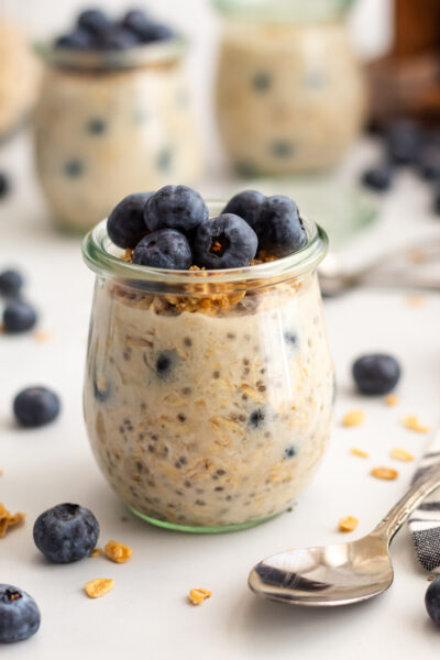 A front facing view of blueberry overnight oats in a clear jar topped with crunchy granola and blueberries, styled on a white surface.