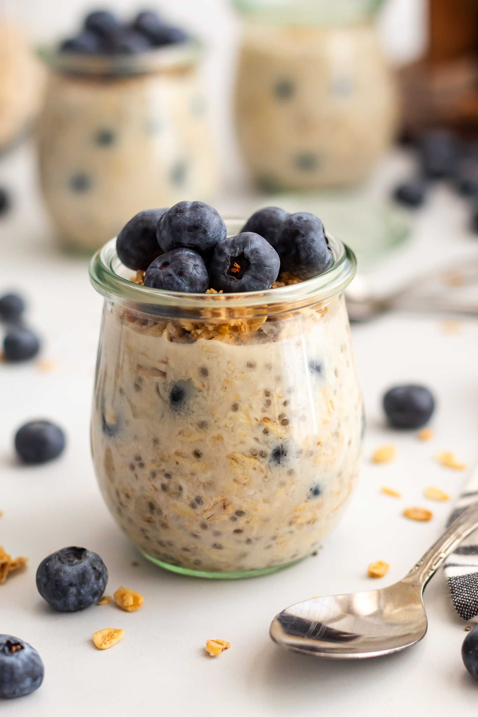A front facing view of blueberry overnight oats in a clear jar topped with crunchy granola and blueberries, styled on a white surface.