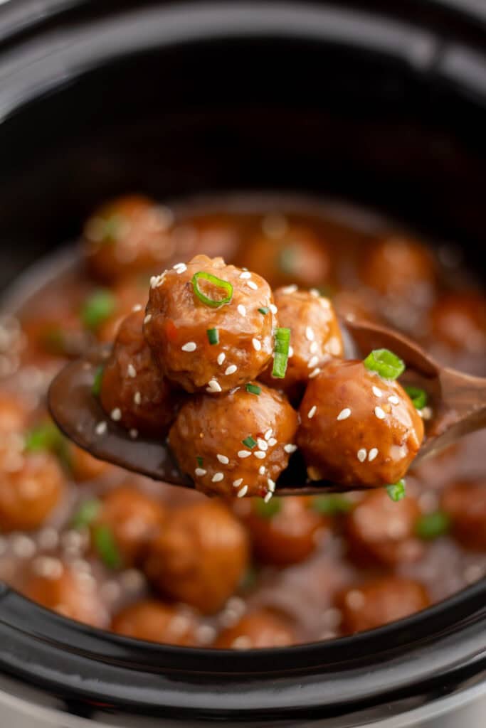 A wooden spoon lifting several crockpot teriyaki meatballs from the slow cooker, showing the rich sauce and sesame seed garnish.