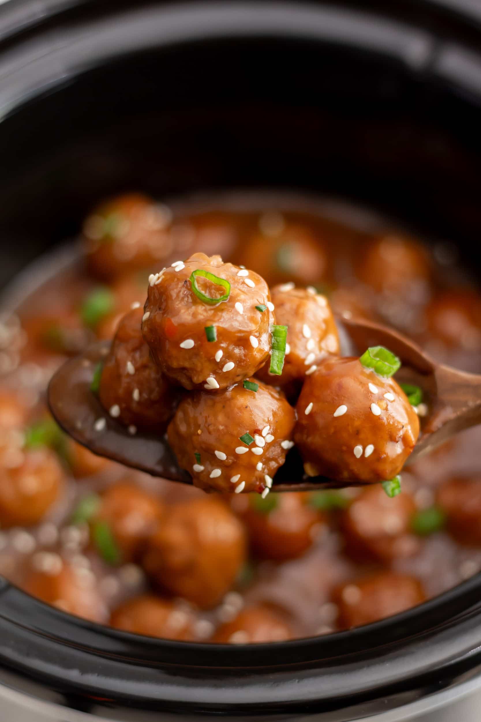 A wooden spoon lifting several crockpot teriyaki meatballs from the slow cooker, showing the rich sauce and sesame seed garnish.