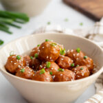 Crockpot teriyaki meatballs served in a cream colored bowl with sauce, garnished with green onions and sesame seeds.