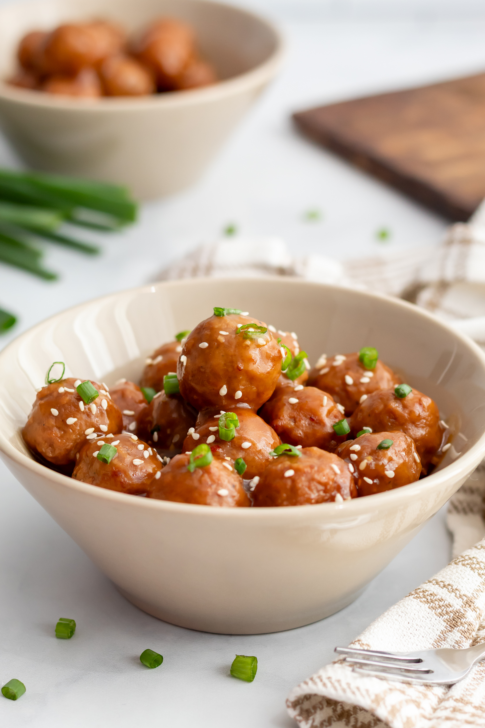 Crockpot teriyaki meatballs served in a cream colored bowl with sauce, garnished with green onions and sesame seeds.
