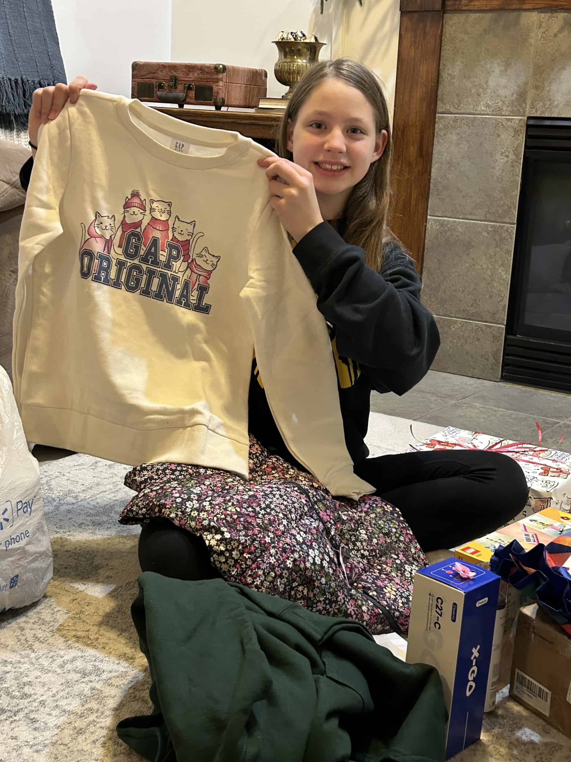 girl smiling and hoilding up a sweatshirt and new clothing that she recevied for her birthday