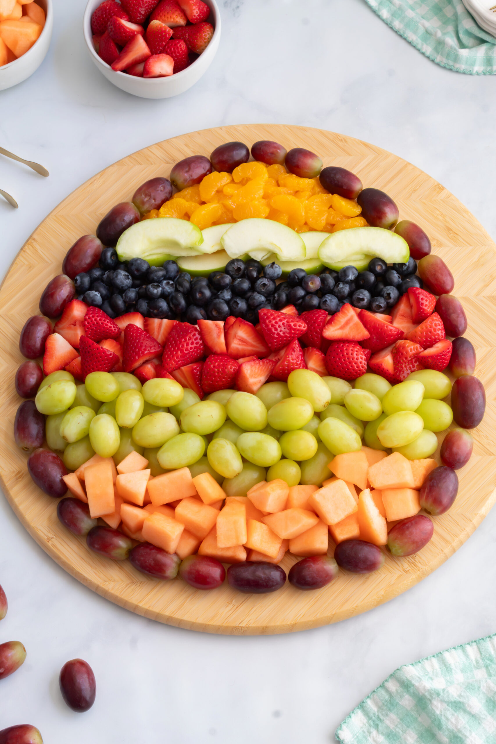 Overhead view of an Easter egg shaped fruit charcuterie board made with red grapes, green grapes, strawberries, blueberries, green apple slices, cantaloupe, and mandarin oranges on a round wooden board