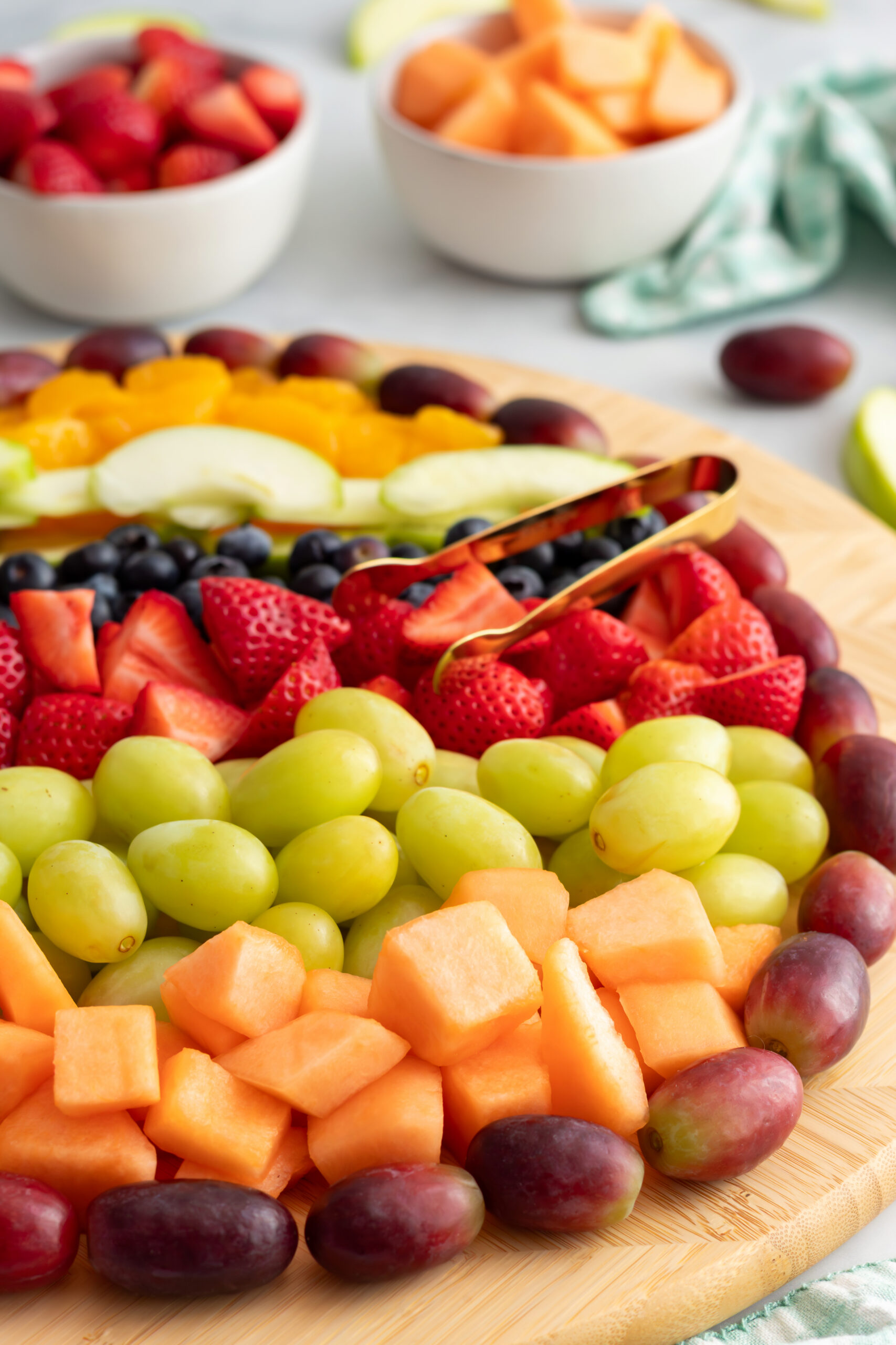 Angled close-up of an Easter egg fruit charcuterie board showing rows of cantaloupe, green grapes, strawberries, and blueberries with gold tongs and a green gingham napkin