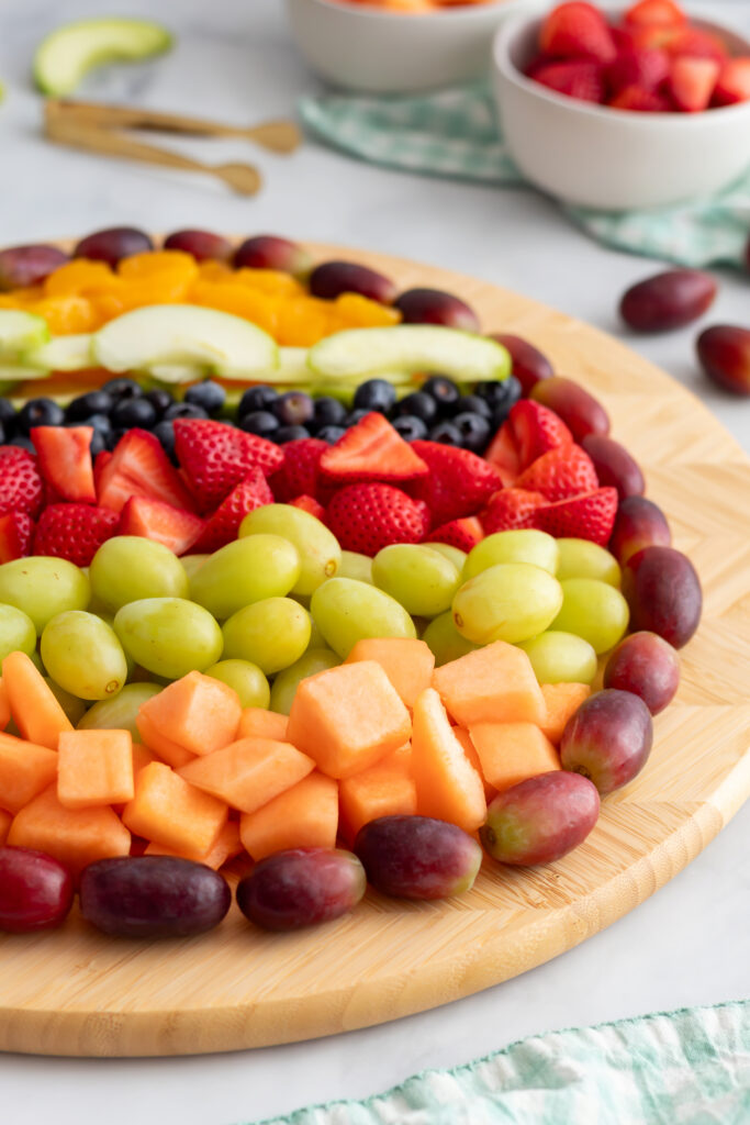 Close-up of colorful fruit stripes on an Easter egg charcuterie board showing rows of cantaloupe, green grapes, strawberries, and blueberries with a soft blurred background