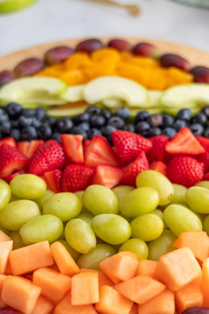 Angled view of the lower half of an Easter egg fruit charcuterie board showing rows of cantaloupe, green grapes, strawberries, and red grape outline with prop bowls in the background