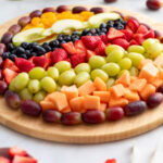 Full view of an Easter egg shaped fruit charcuterie board on a round wooden board with scattered strawberries, green apple slices, and gold tongs alongside