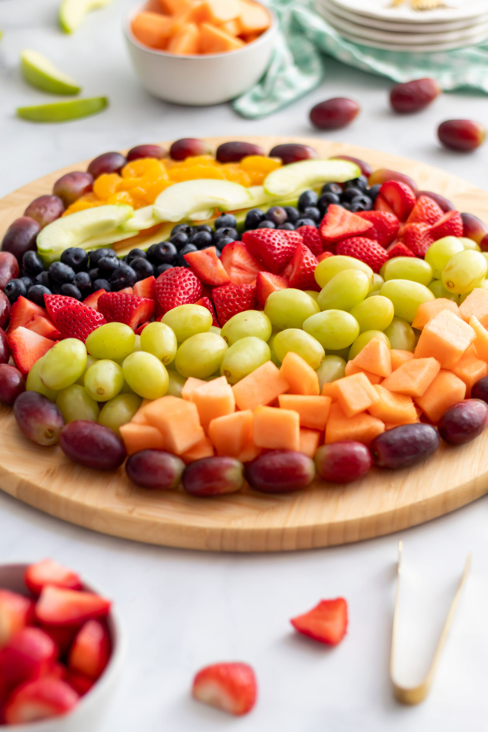 Full view of an Easter egg shaped fruit charcuterie board on a round wooden board with scattered strawberries, green apple slices, and gold tongs alongside