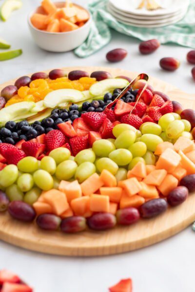 Easter egg fruit charcuterie board on a round wooden board with gold tongs resting across the fruit and white bowls and green gingham napkin in the background