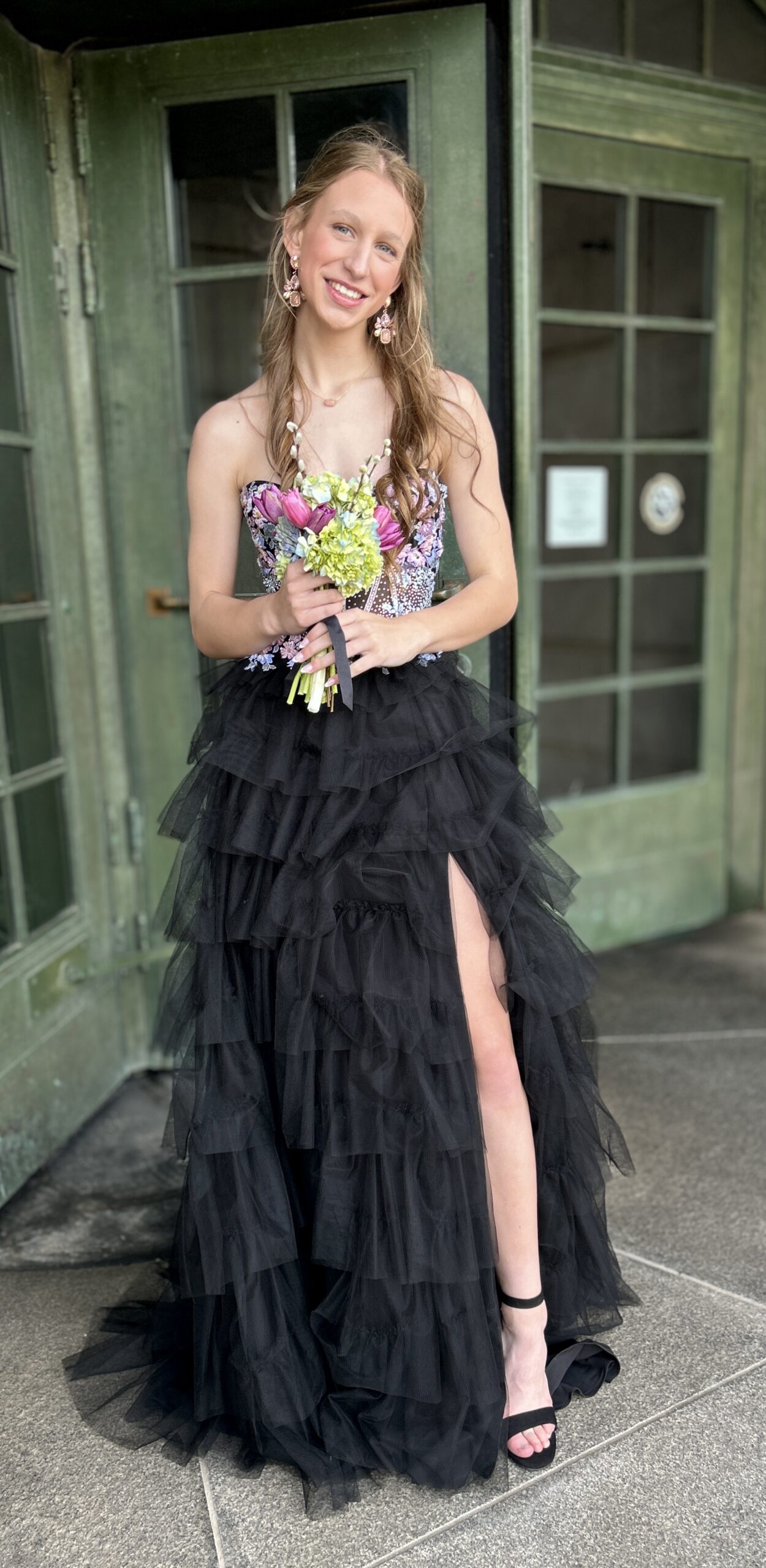Girl in black prom dress with ruffled bottom and beaded top holding a bouquet of spring flowers and standing and smiling for photos