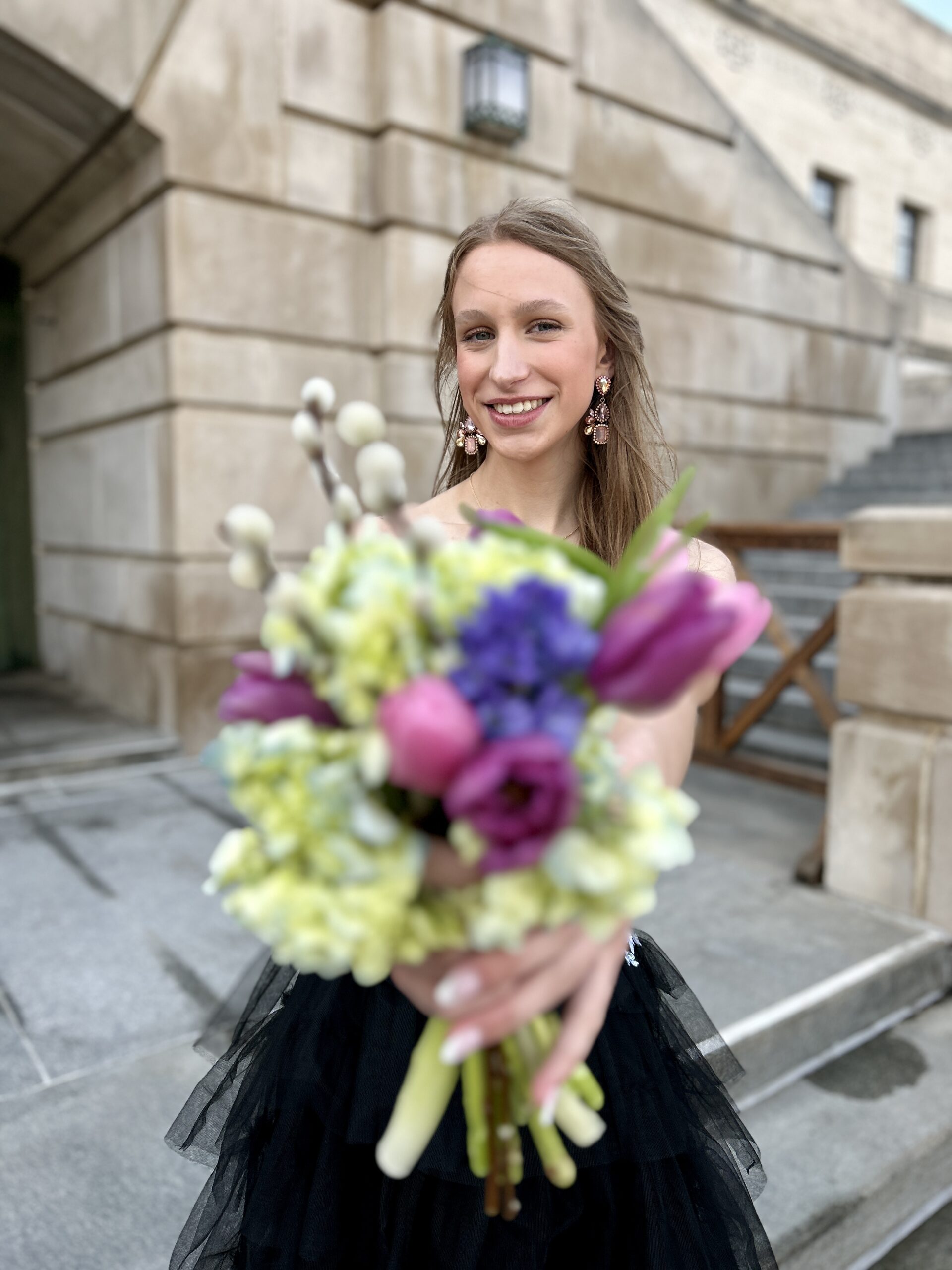 Girl in black prom dress with ruffled bottom and beaded top holding a bouquet of spring flowers and standing and smiling for photos