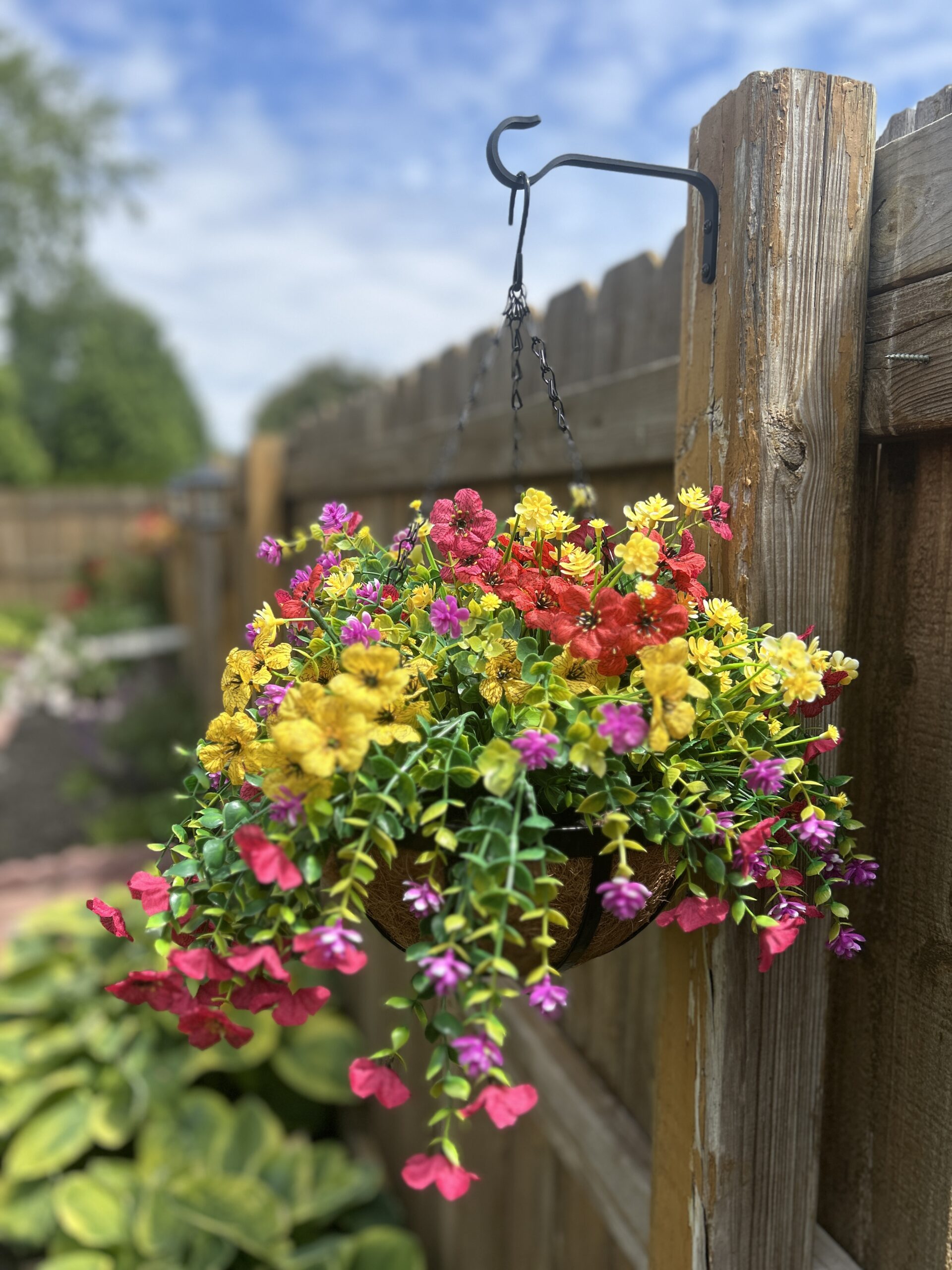 Hanging basket hanging on a hook from a wooden fence outdoors filled with colorful faux silk flowers