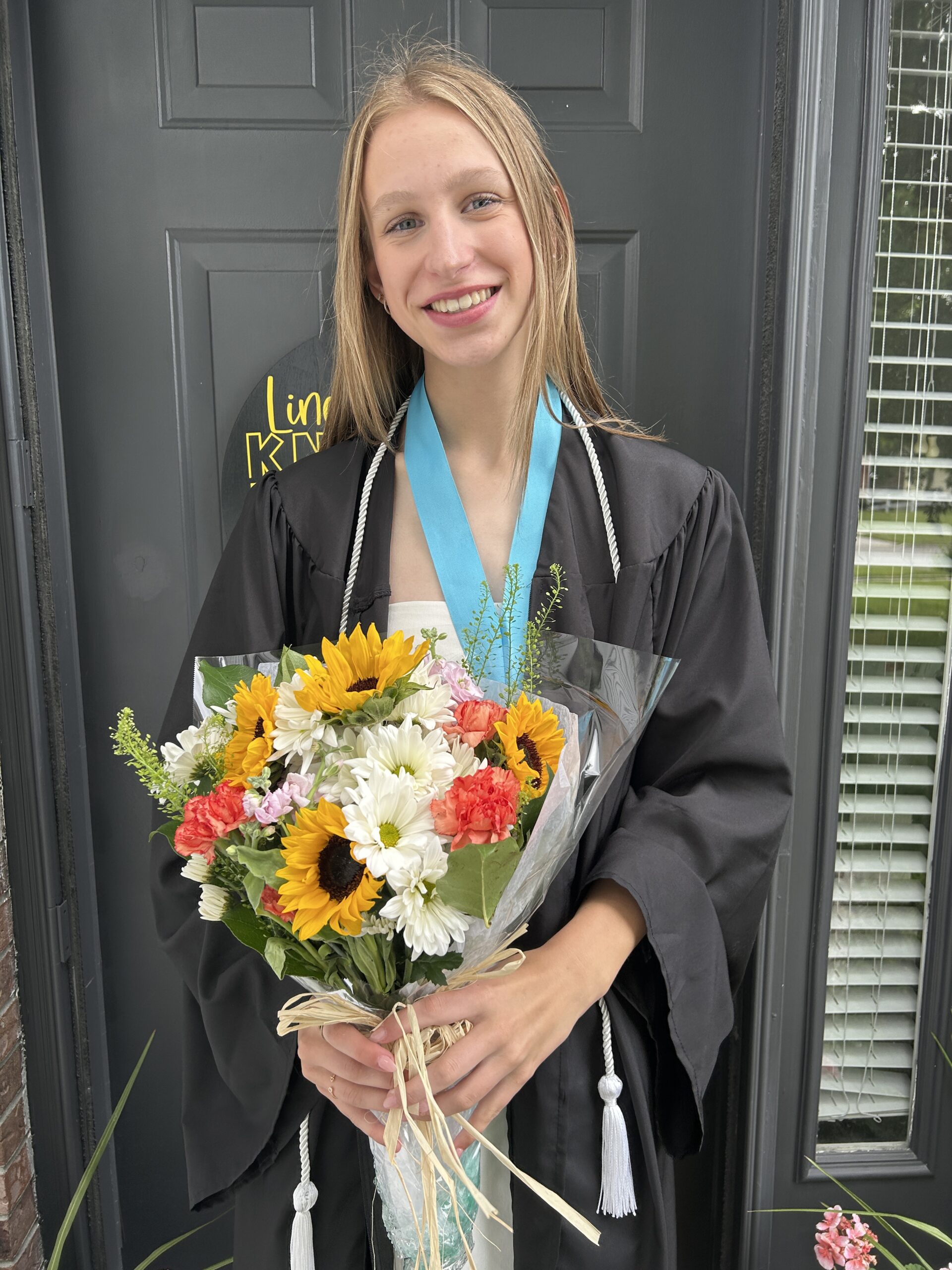 Girl standing on the porch in her graduation gown holding a brightly colored bouquet of flowers