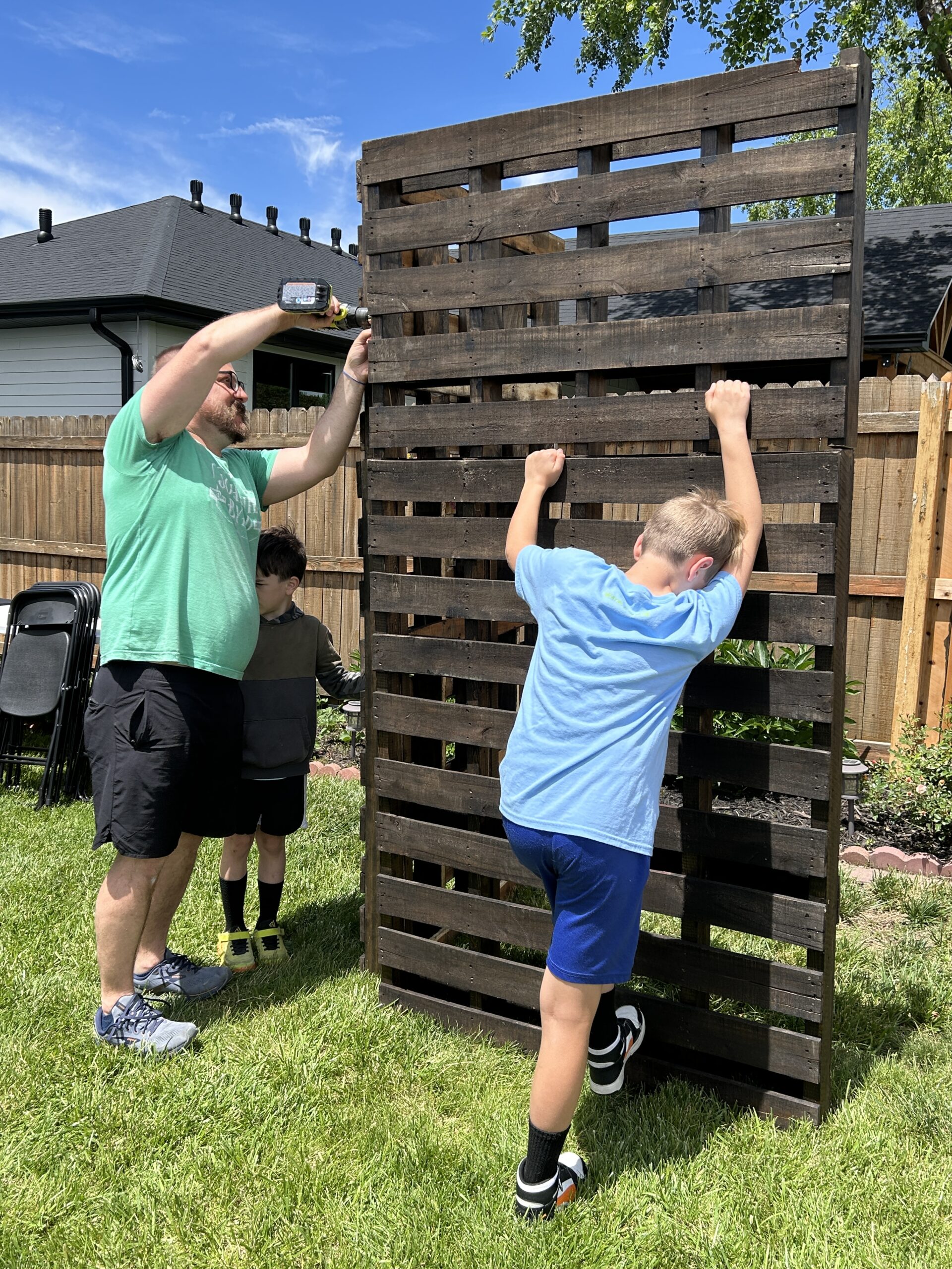 Man holding a drill and building a pallet backdrop while a young boy helps hold up the pieces