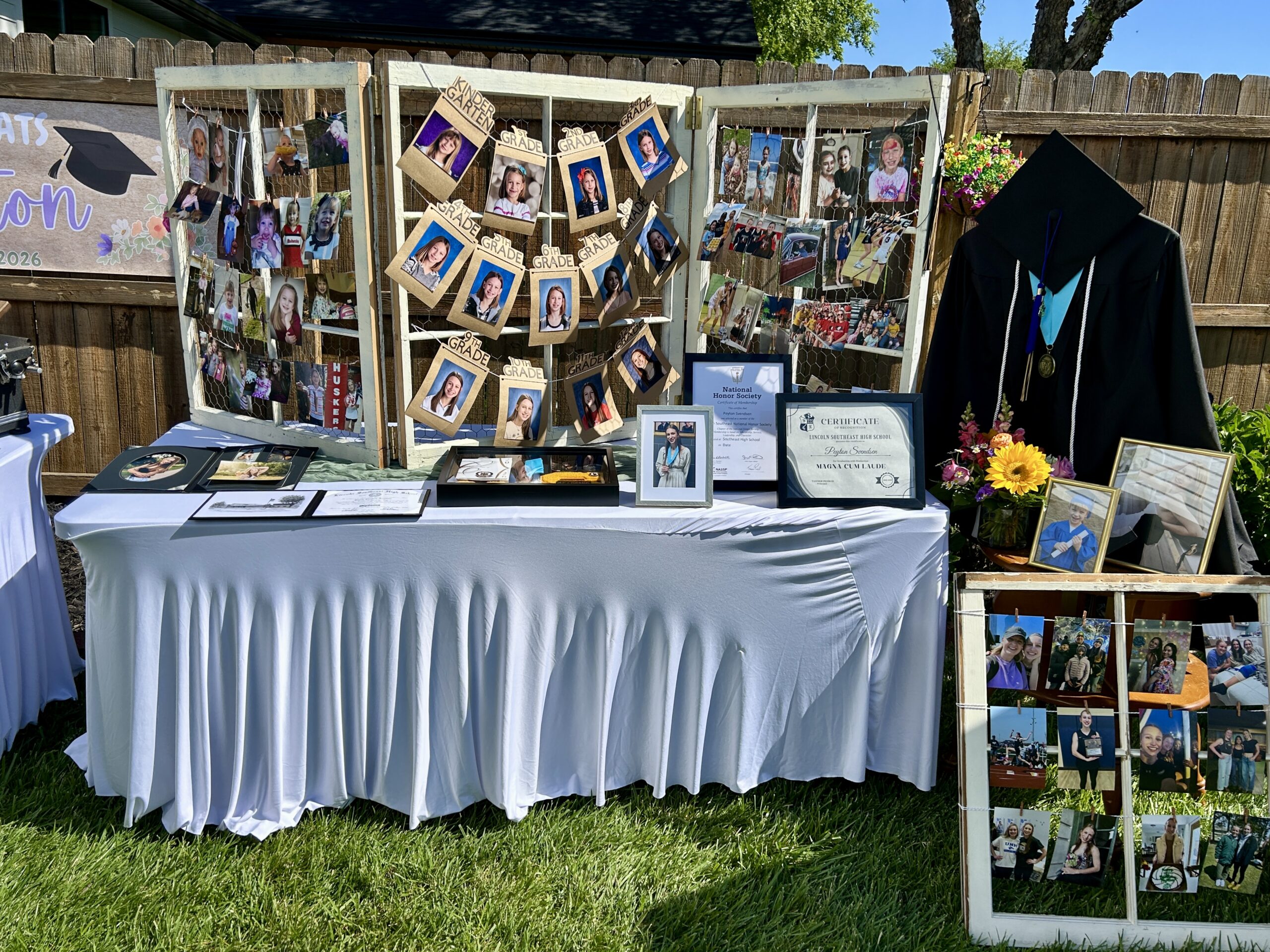 High school graduation party display featuring childhood photos, senior portraits, awards, diploma, and graduation cap on an outdoor memory table.