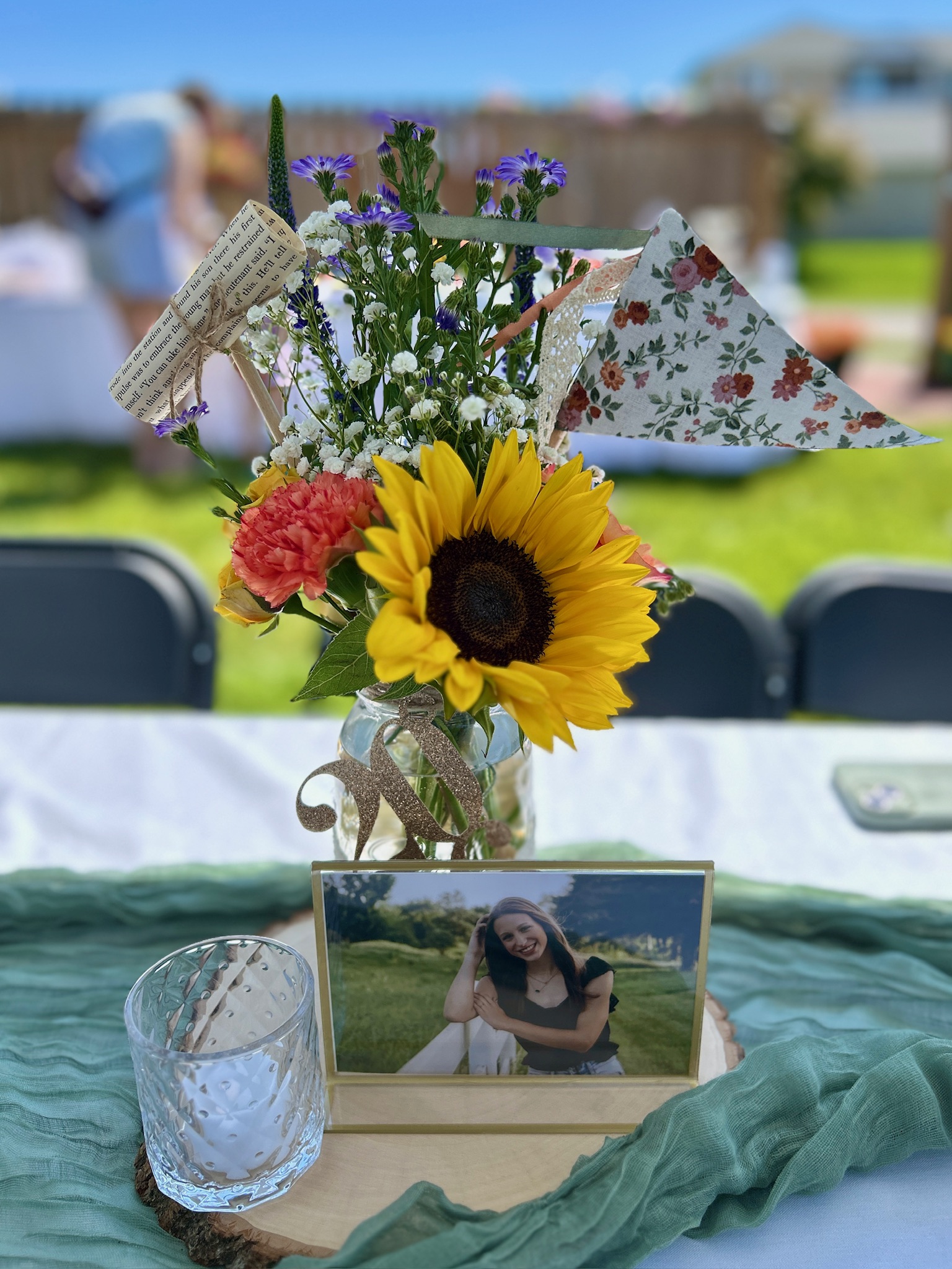 Floral graduation party centerpiece in a ball jar with sunflower, baby's breath, wildflowers, vintage-inspired pennant banner, bookpage diploma, votive candle holder, and framed senior photo.