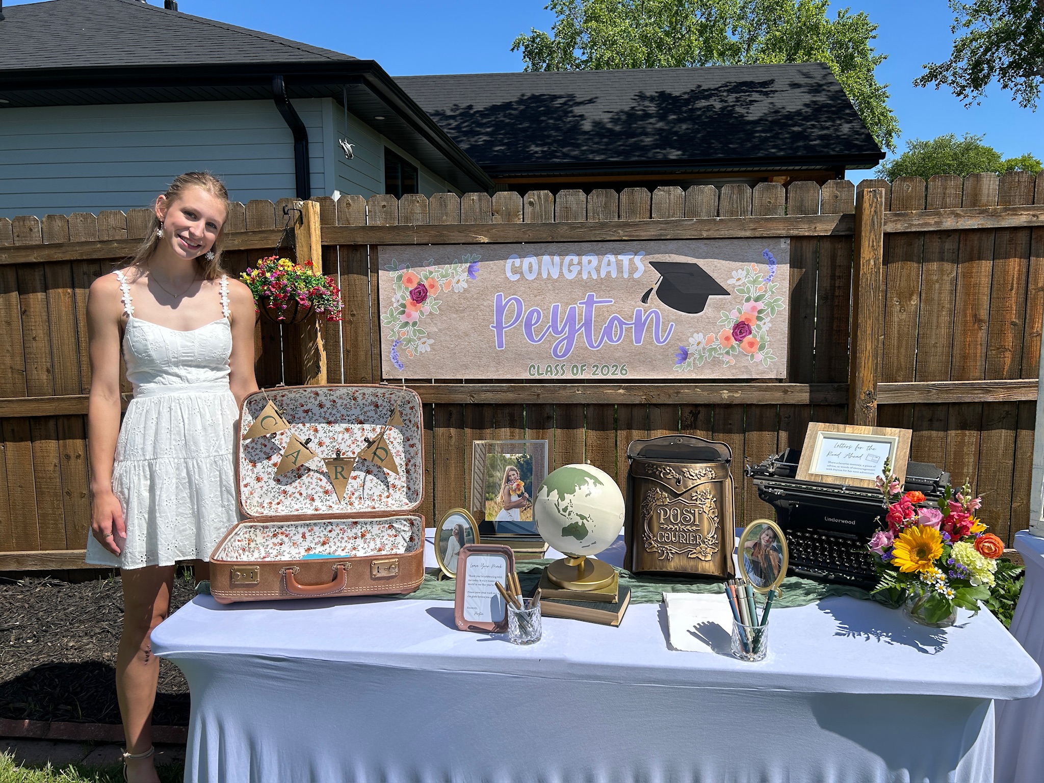 Girl standing and smiling behind the guest book table at her graduation party. There is is congratulations banner on the back fence and the table is filled with vintage decor including a suitecase card box and diy globe guest book. There is also an old post box for notes to the grad, a vintage typewriter, a bouqet of colorful flowers, and framed photos fo the graduate.