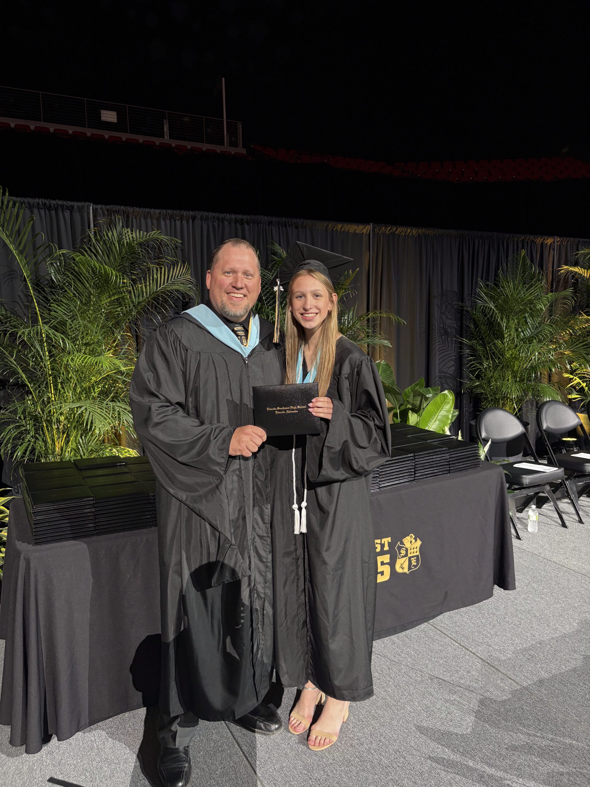 Dad and daughter in graduation regalia posing for a pohto while he hands her a dipoloma