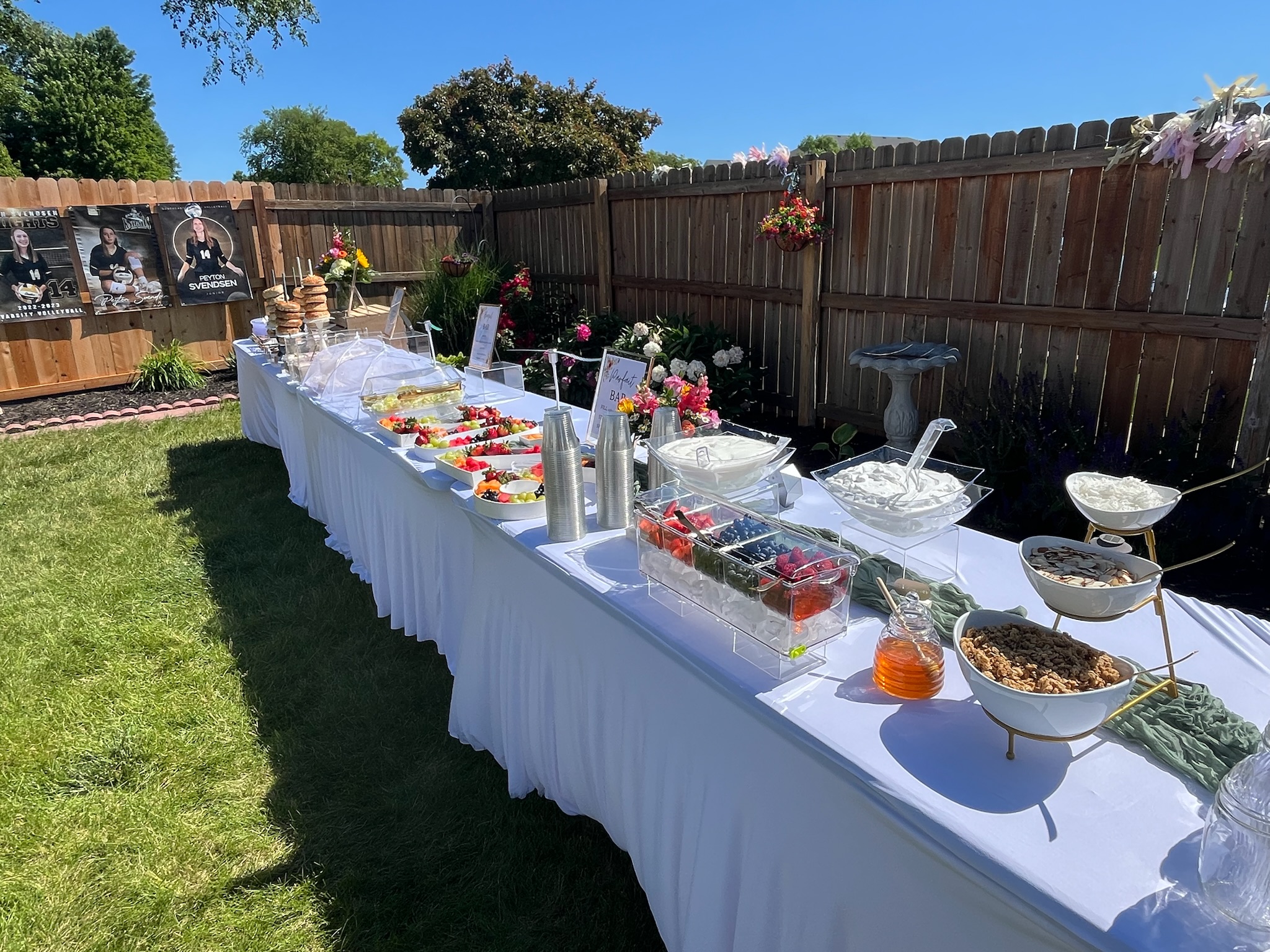 Outdoor graduation brunch buffet featuring a yogurt parfait bar, fresh fruit display, bagel station, and floral decorations.