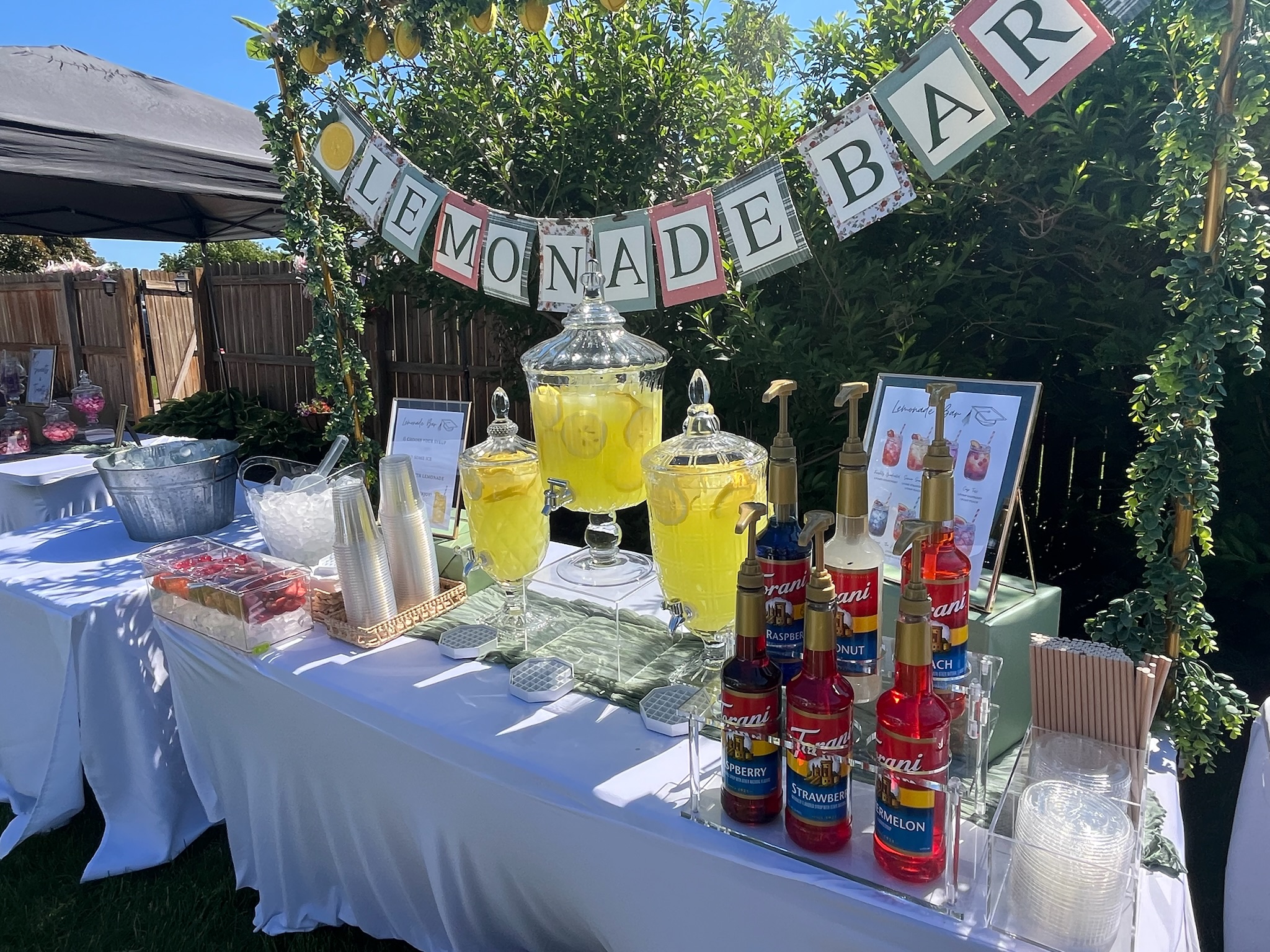 Graduation party lemonade bar featuring flavored syrups, fresh lemonade dispensers, decorative lemon arch, and custom signage.