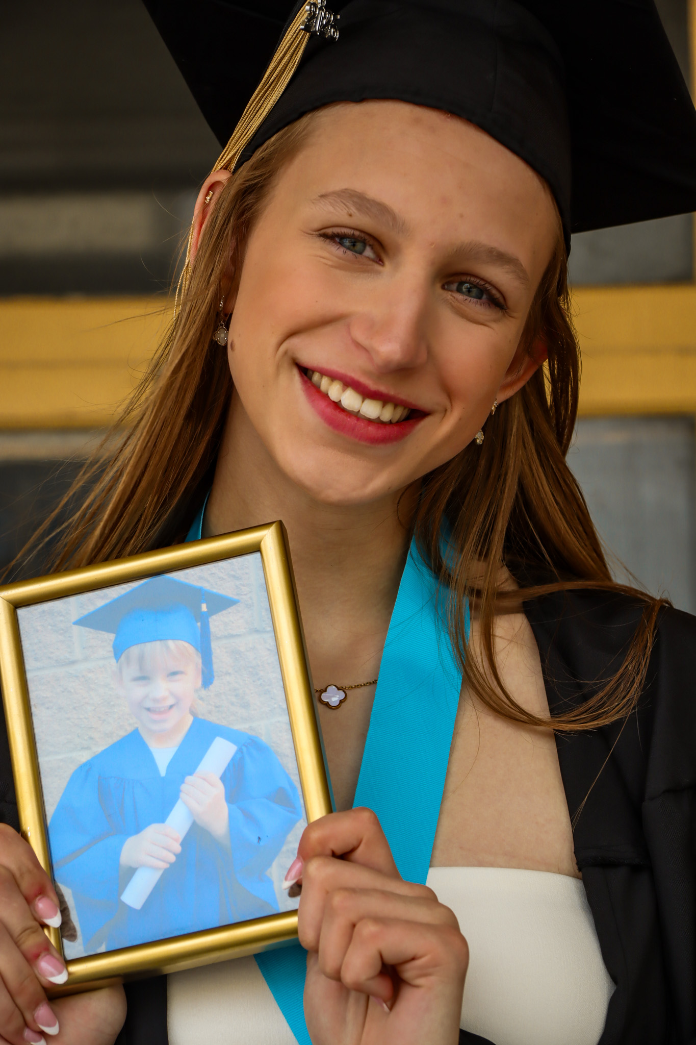 Girl in cap and gown smiling and holding photo of younger self in pre-school graduation regalia