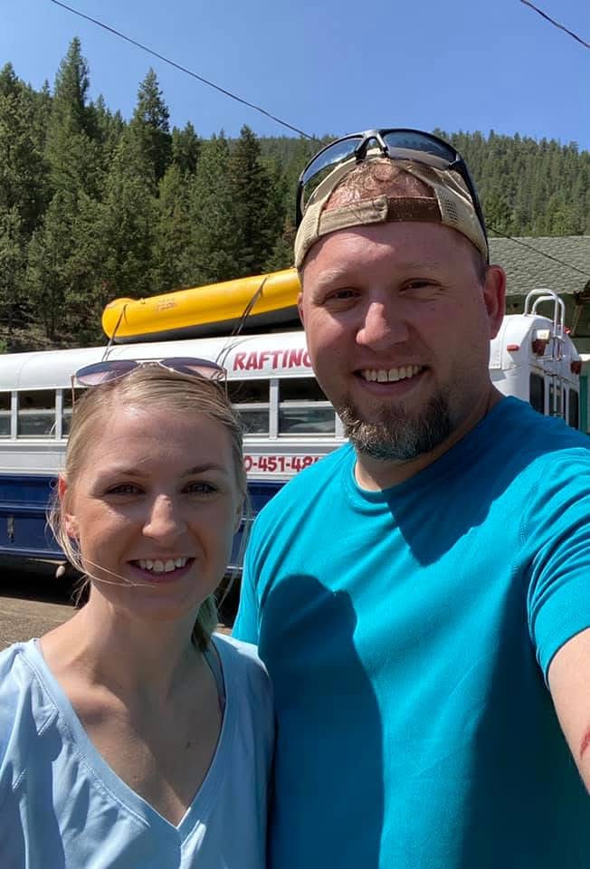 Couple smiling on a tripto colorado with a forest of trees in the background and a rafting bus with a raft tied on top