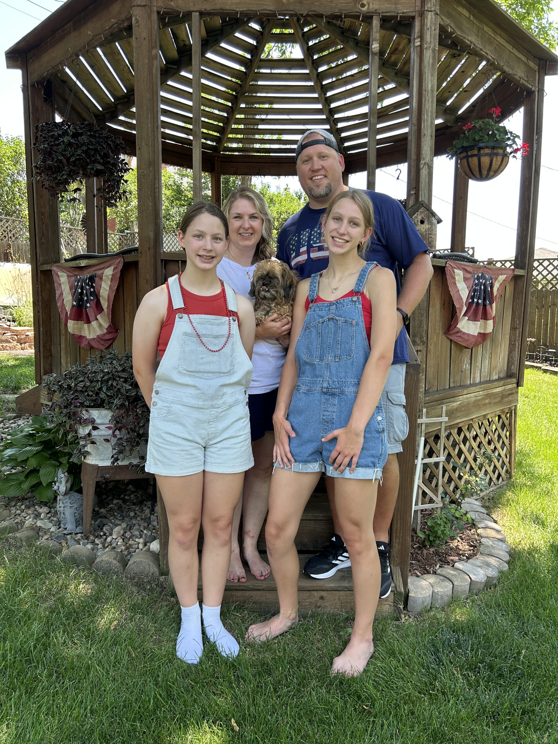 Family dressed in red, white and blue for the 4th of July standing in front of a gazebo smiling for a photo