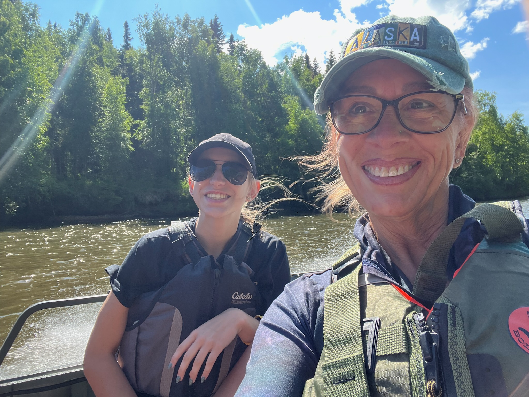 A woman and girl wearing life jackets smile for a selfie while riding in a river boat surrounded by trees in Alaska.