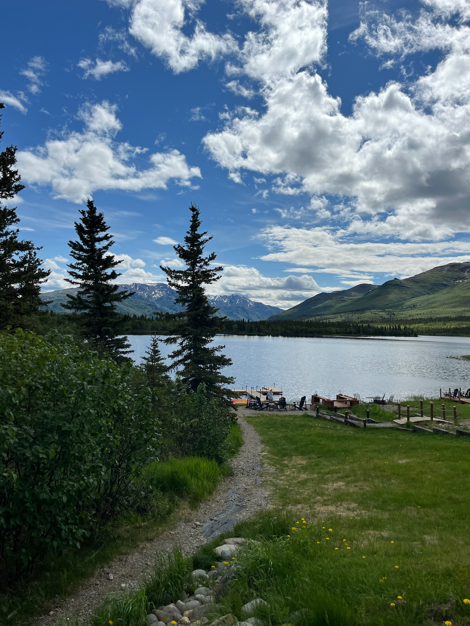 A peaceful mountain lake surrounded by evergreen trees and snow-capped peaks under a blue sky with scattered clouds.