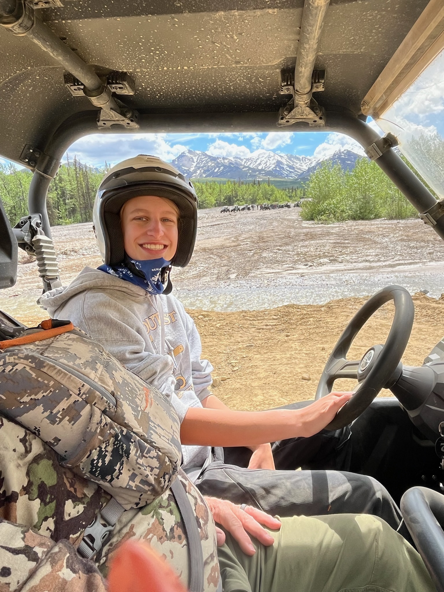 A smiling girl wearing a helmet sits behind the wheel of a side-by-side ATV with snow-capped mountains and a river in the background.