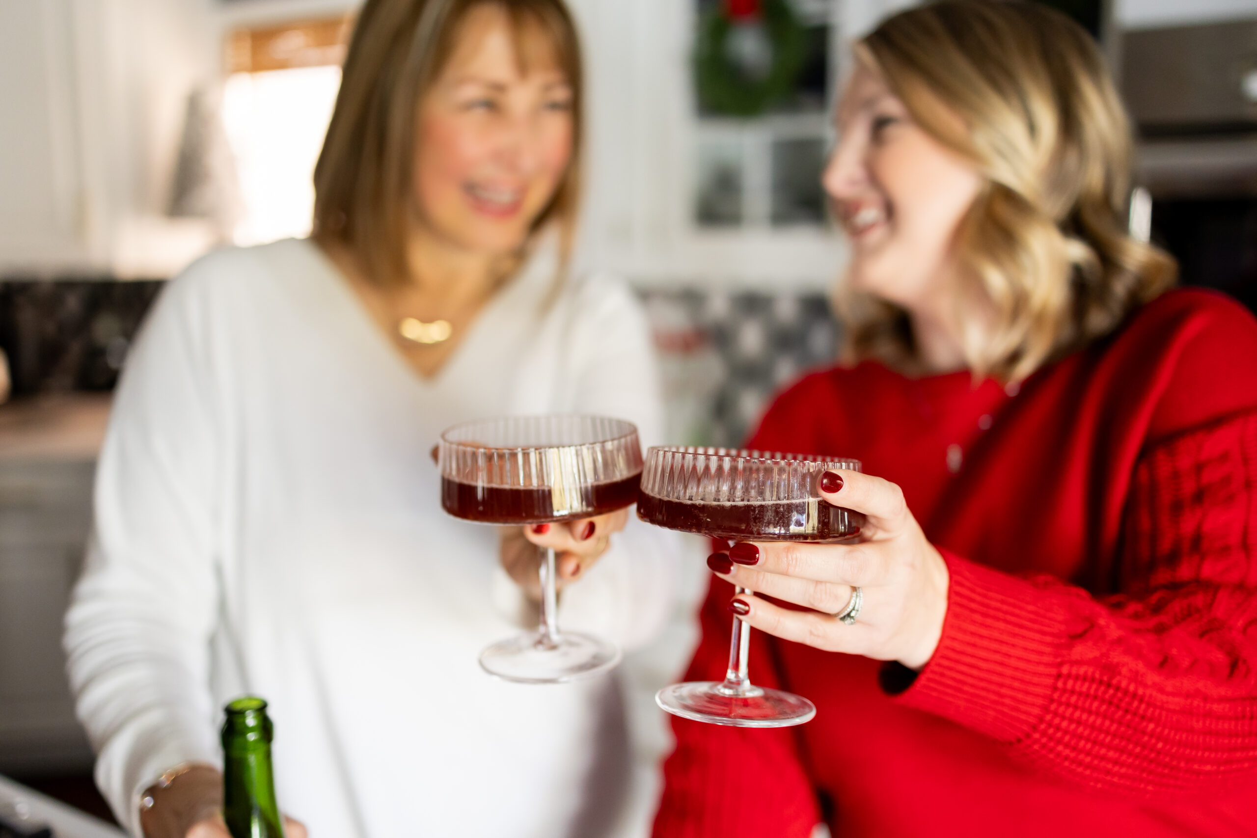 Two women toast with coupe glasses filled with a red holiday cocktail while wearing red and white sweaters.