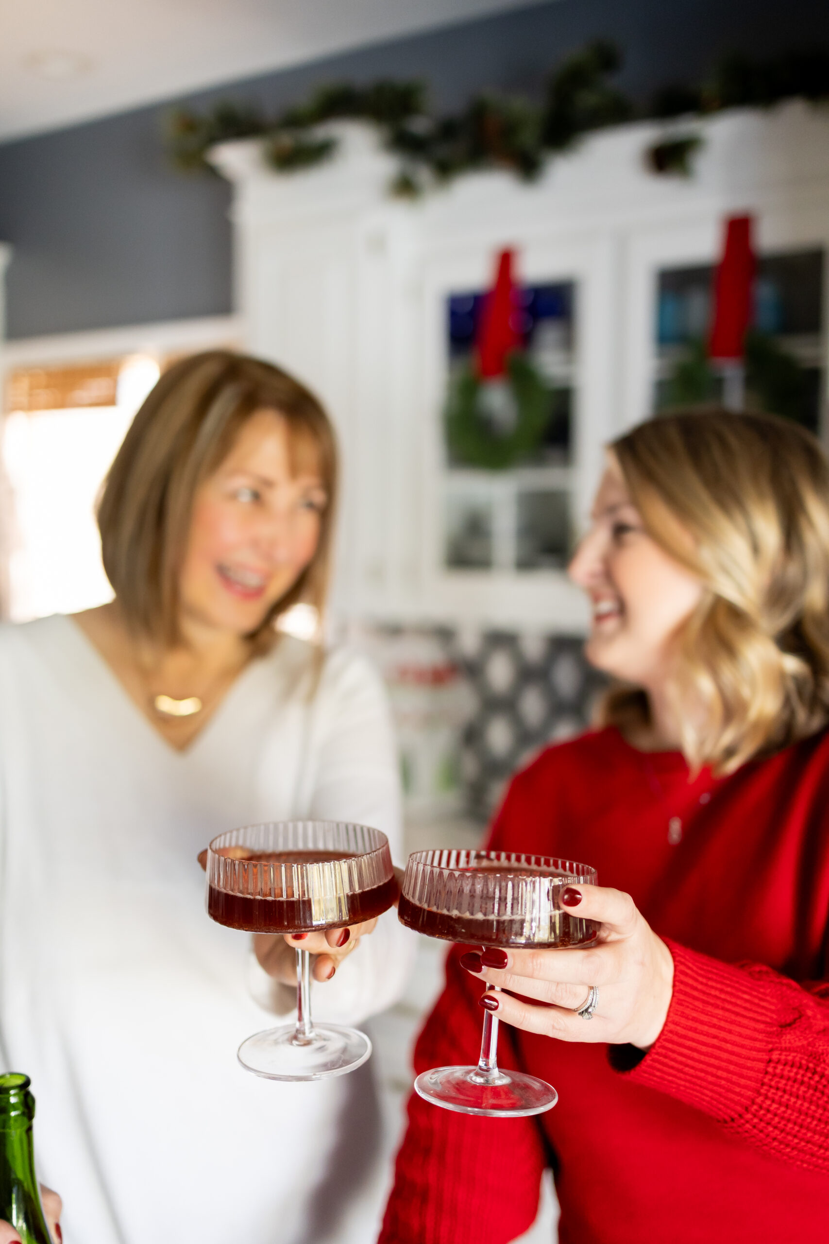 Blurred image of Missy Noonan and Jen Svendsen holding holiday cocktails and toasting in a Christmas-decorated kitchen during their Twinkling Lights & Festive Bites holiday gathering.