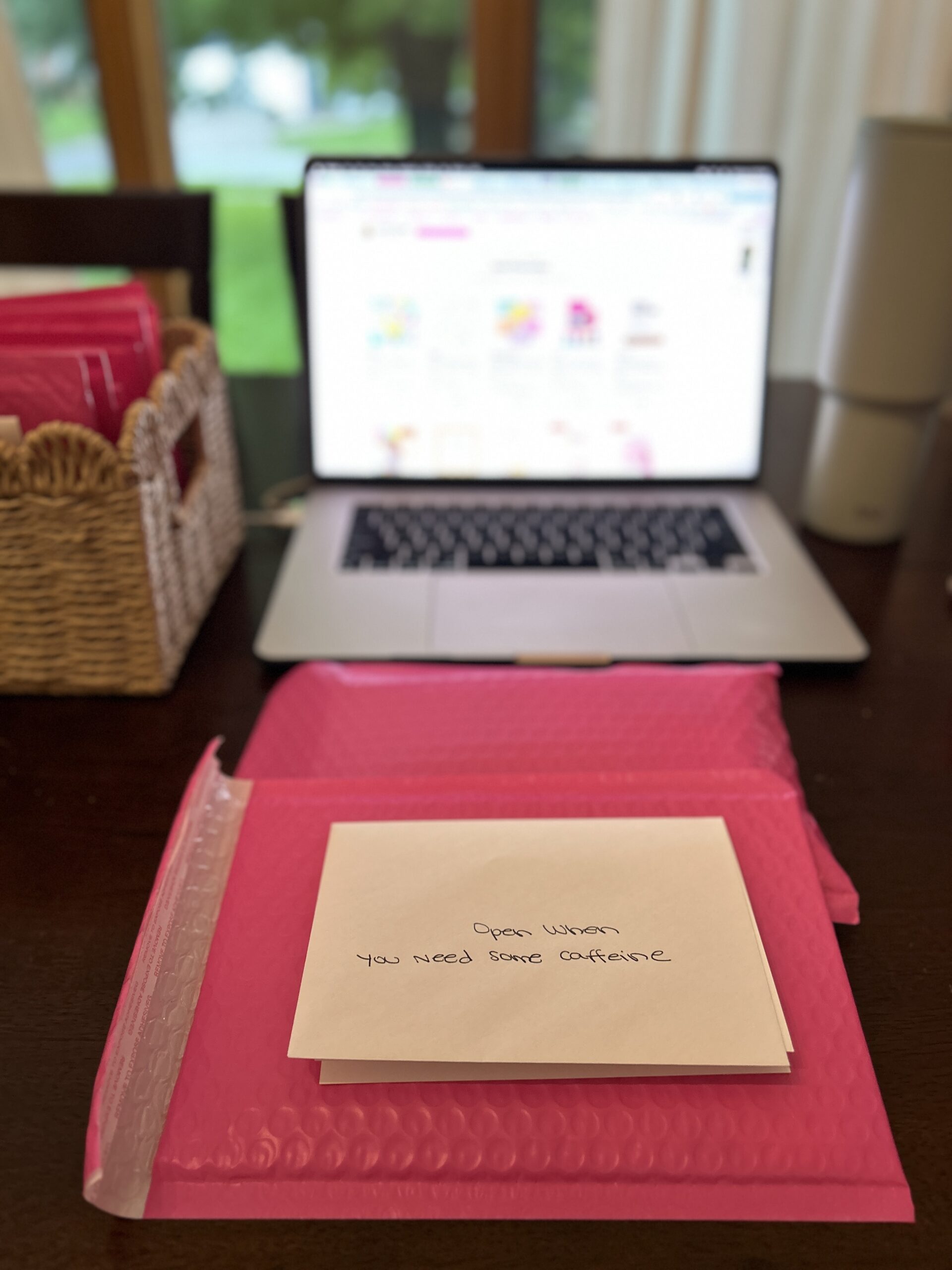 Pink care package with an envelope labeled “Open when you need some caffeine” sitting in front of a laptop.