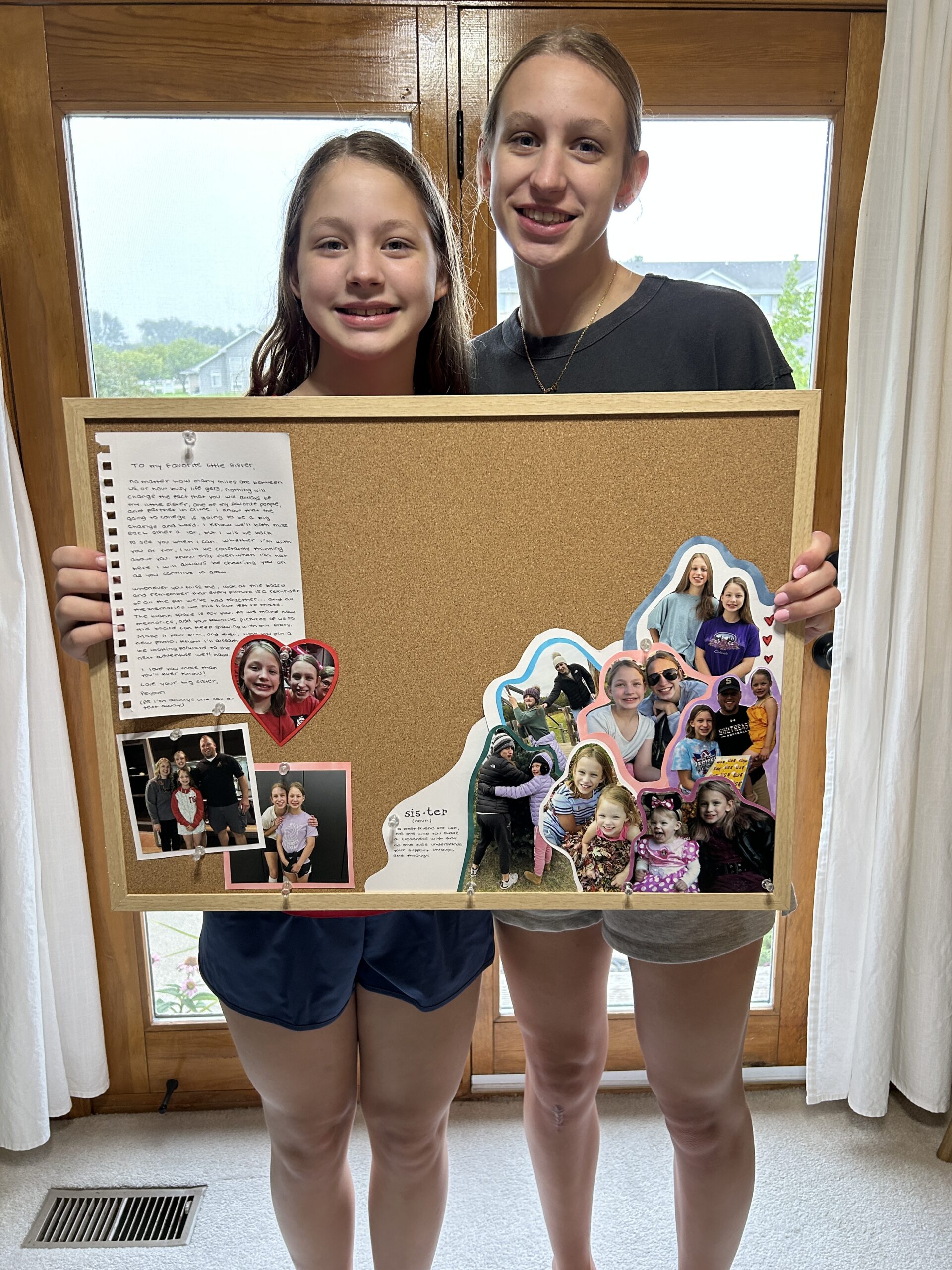 Two sisters hold a handmade corkboard filled with family photos and a handwritten letter before college move-in.