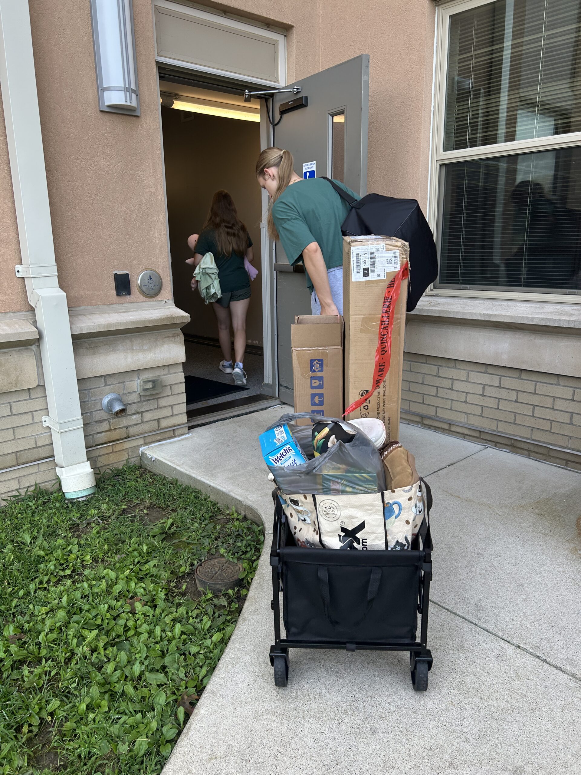 College students carry boxes and supplies into their apartment during move-in day using a collapsible wagon.