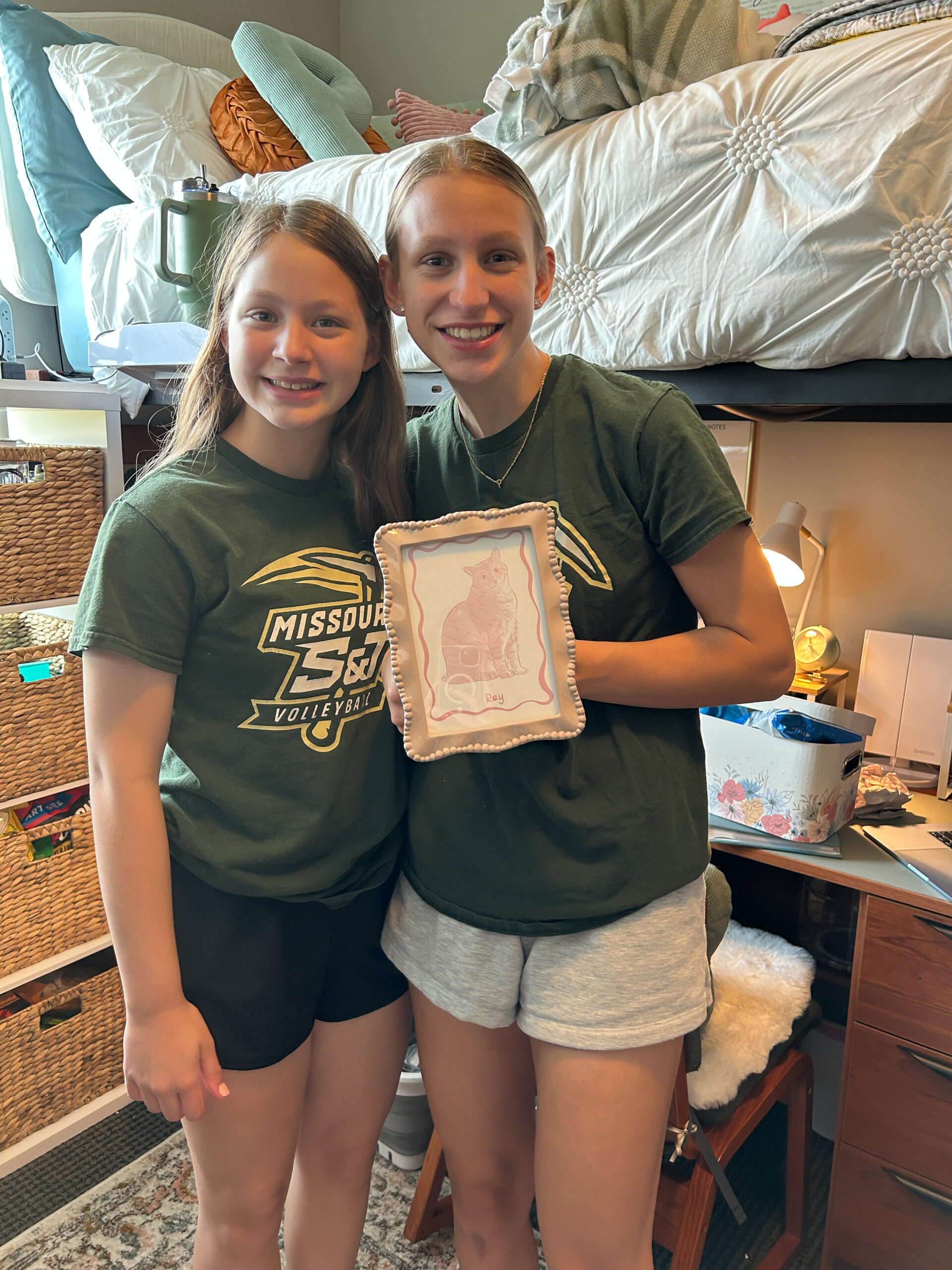 Two sisters wearing matching Missouri S&T volleyball shirts and holding a framed cat illustration in a college bedroom.